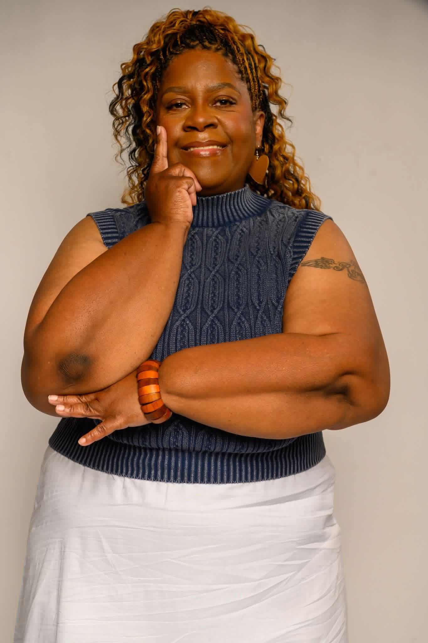 Woman with curly hair wearing a sleeveless blue sweater and white skirt, smiling with her hand on her chin.