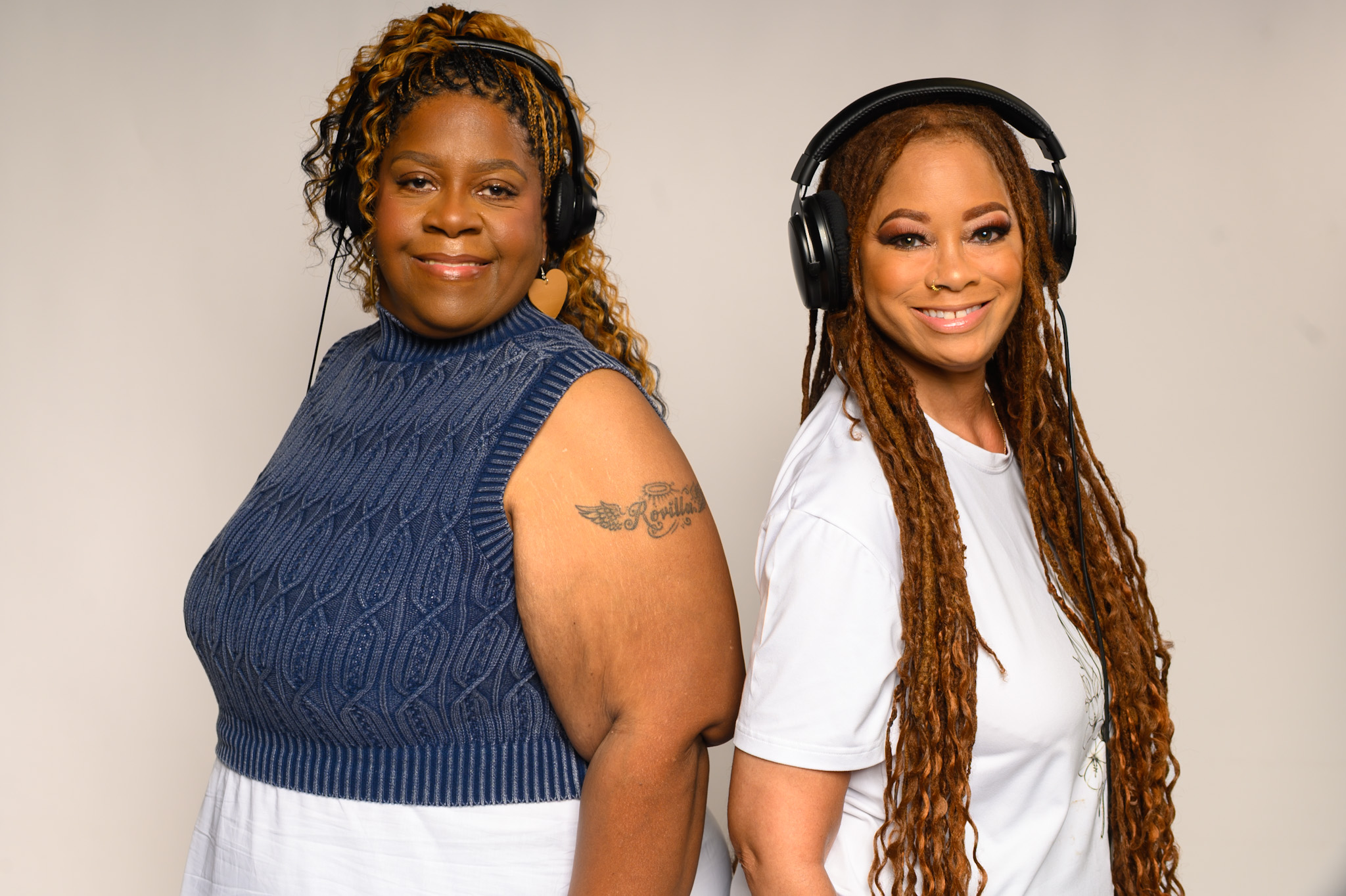 Two women smiling, one with curly hair and a tattoo, the other with long dreadlocks and headphones, standing against a plain background.
