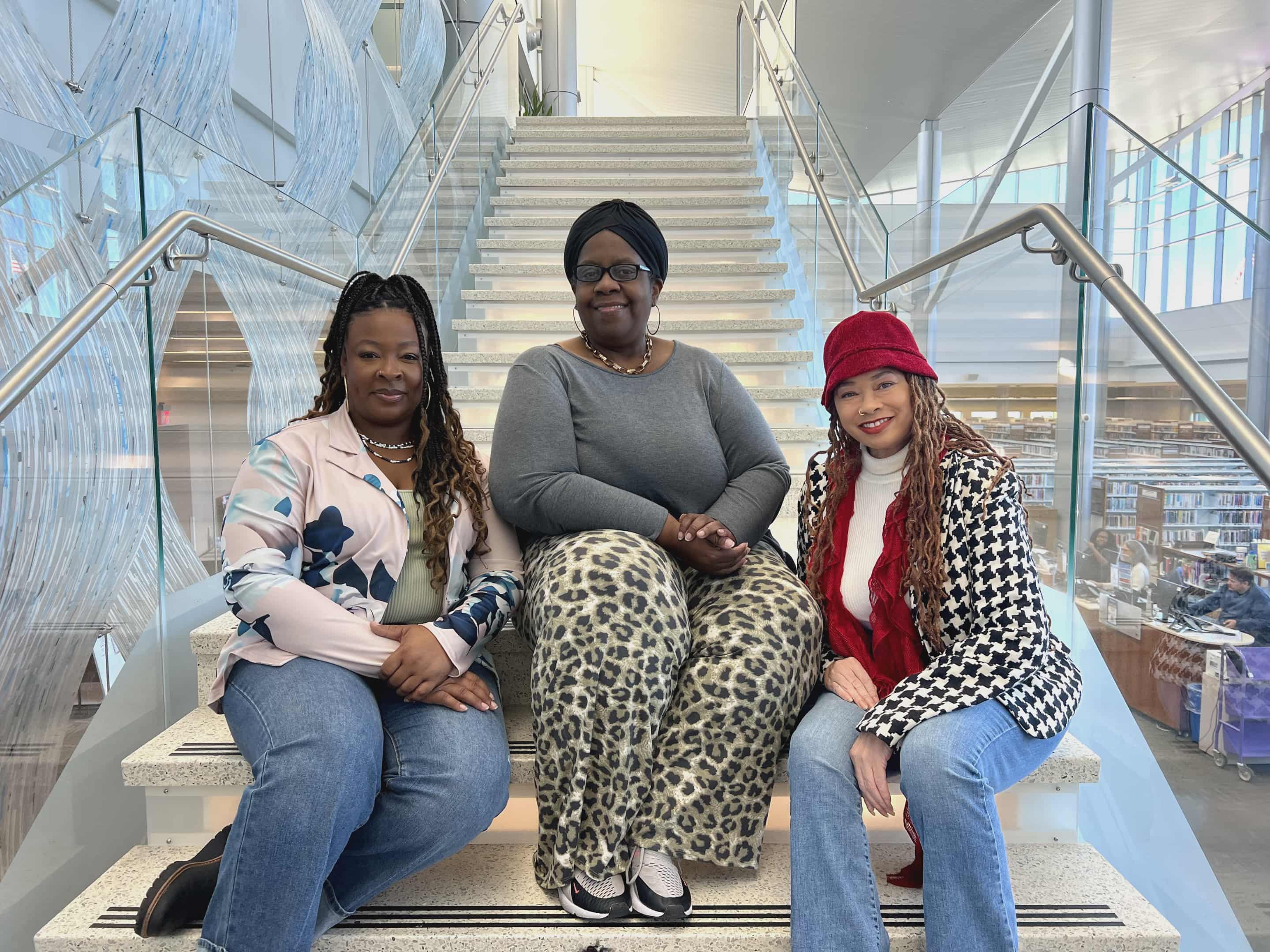 Three women sitting on a staircase indoors, smiling, with glass walls and a shopping area in the background.