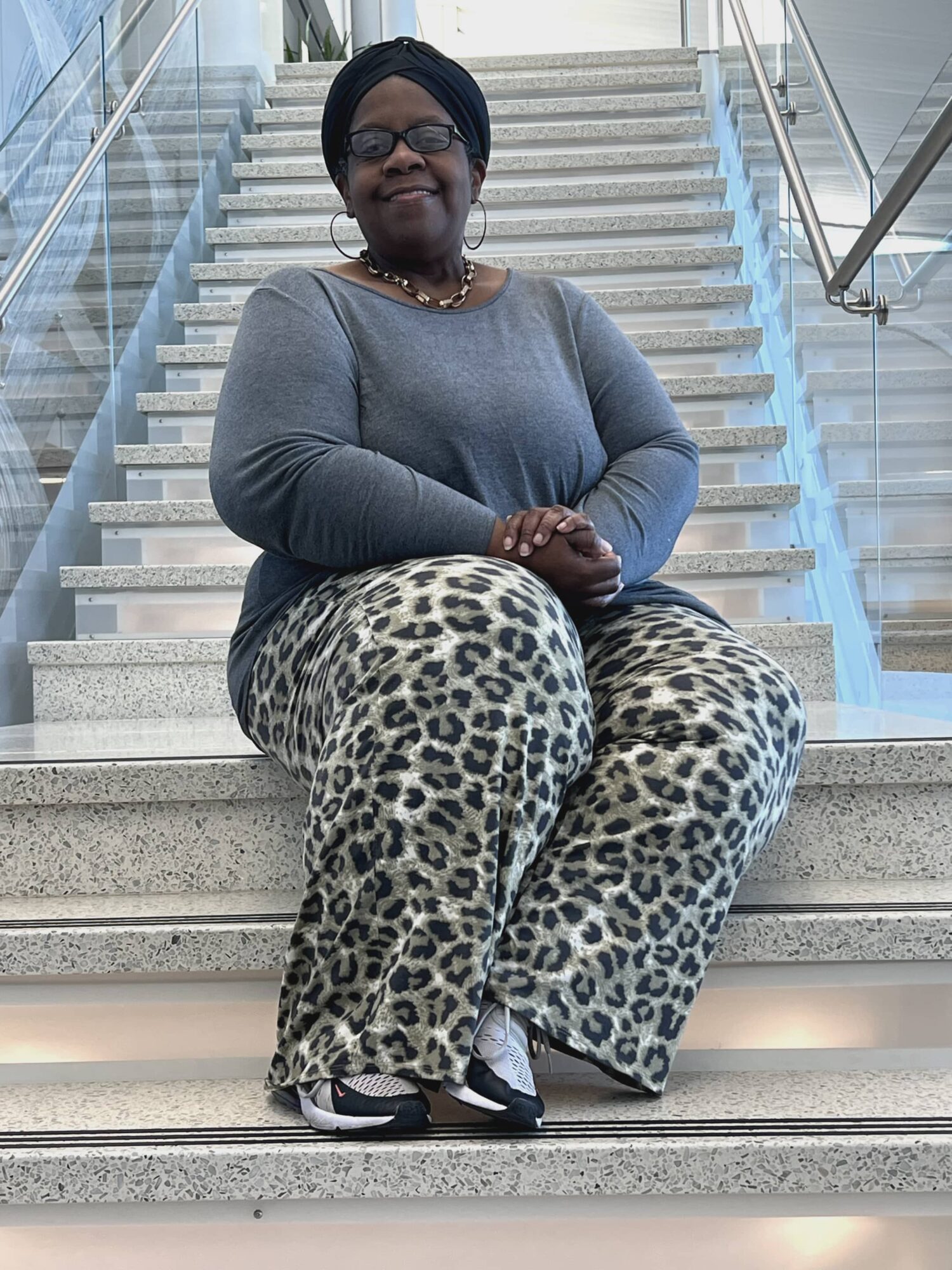 Woman sitting on stairs, wearing glasses, a headscarf, a gray top, and leopard print pants, smiling at camera.