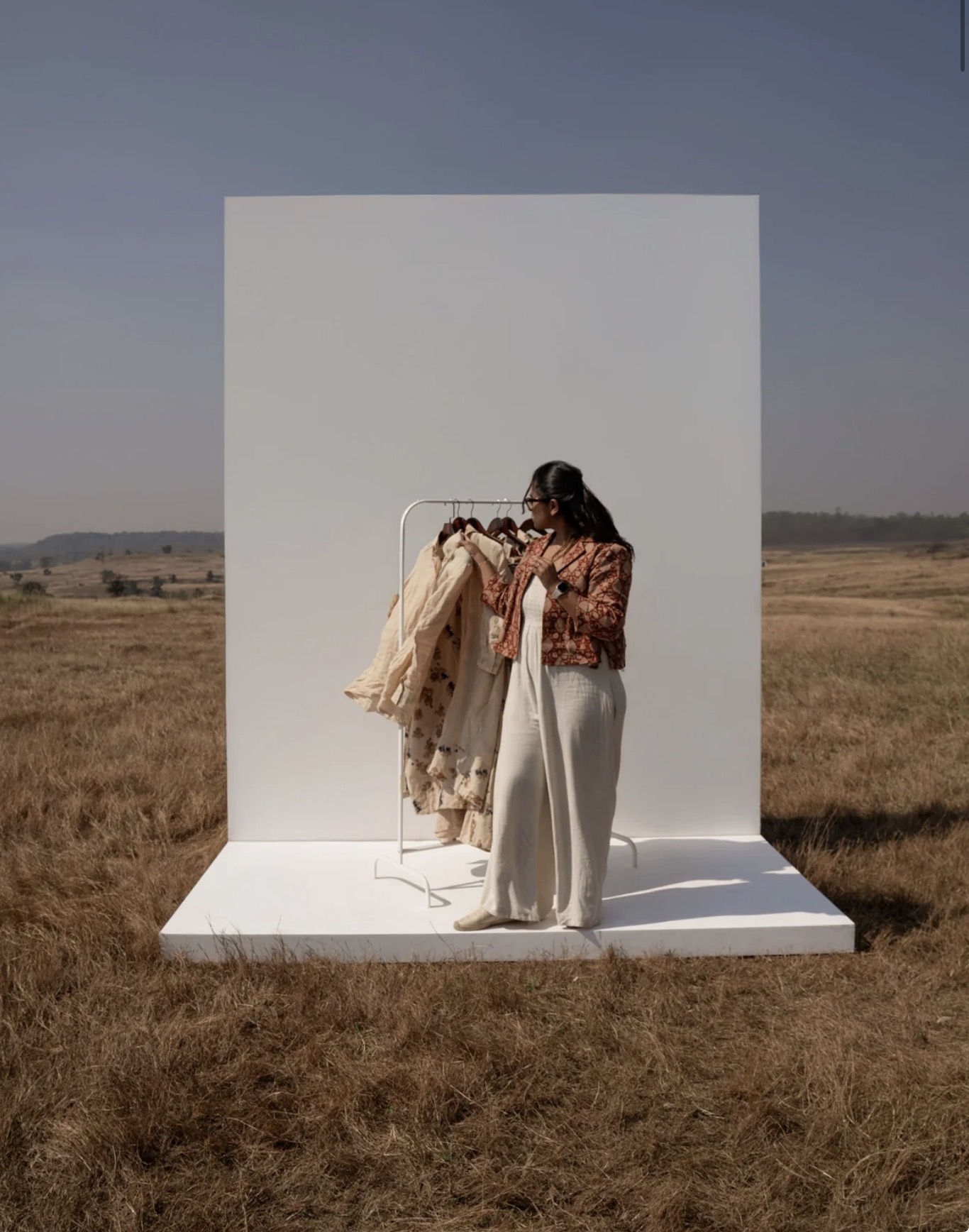 Woman with sunglasses stands next to clothing rack outdoors with a white backdrop, in a grassy field under a clear sky.