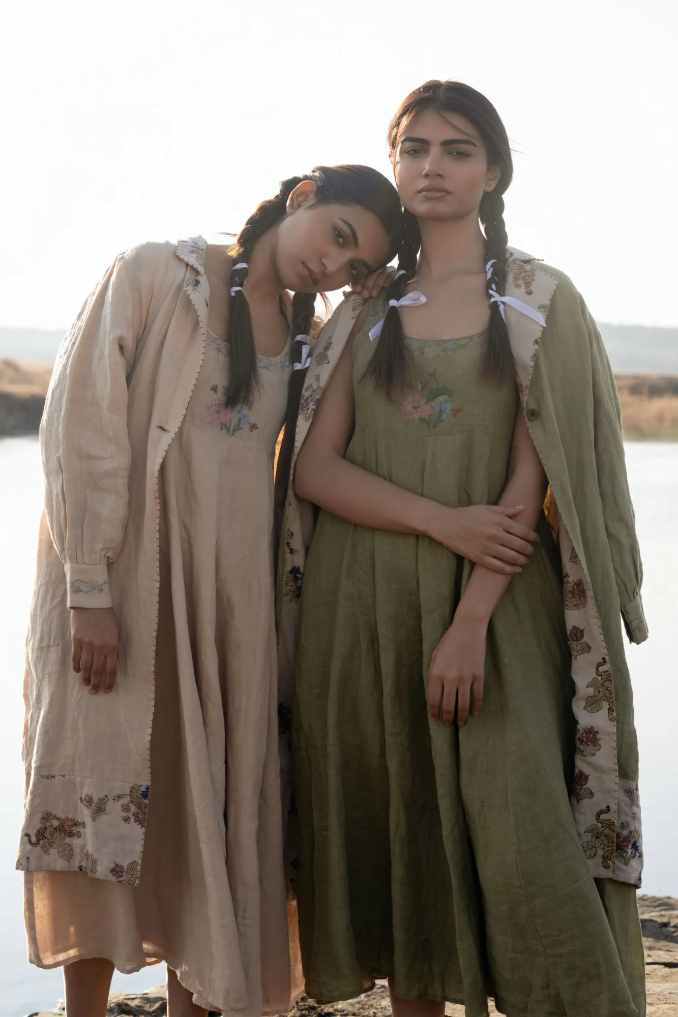 Two women stand outdoors, one resting her head on the other's shoulder, both wearing long dresses and jackets.
