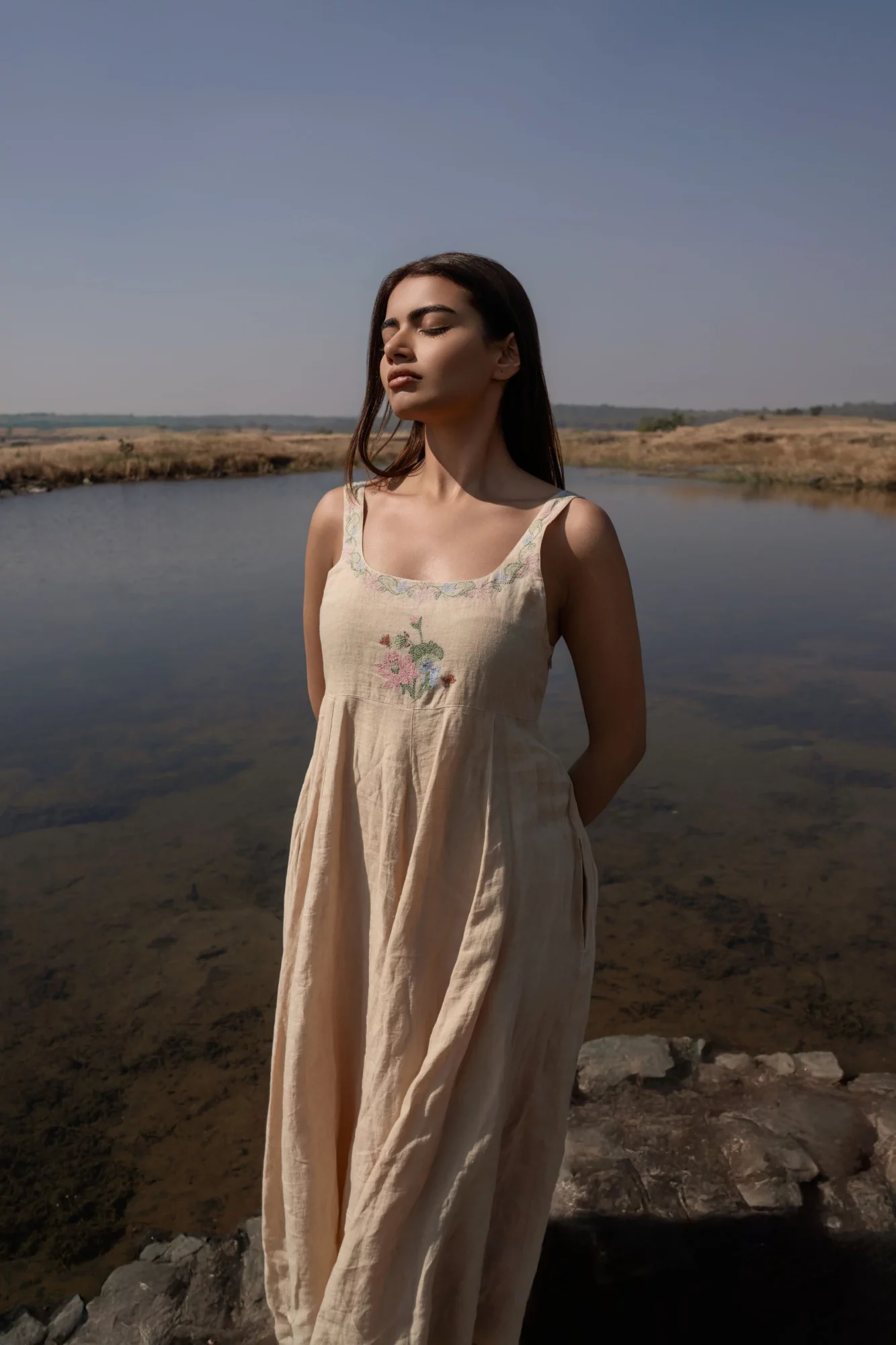 Young woman in a light-colored dress standing outdoors near water, with a landscape background and clear sky.