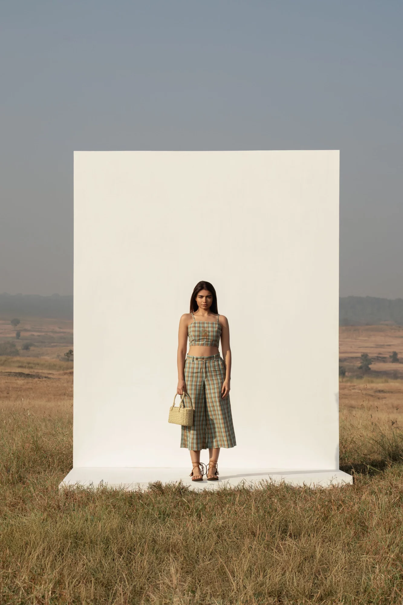 Woman standing in front of a white rectangular backdrop in an open field, holding a bag, wearing a checkered dress.
