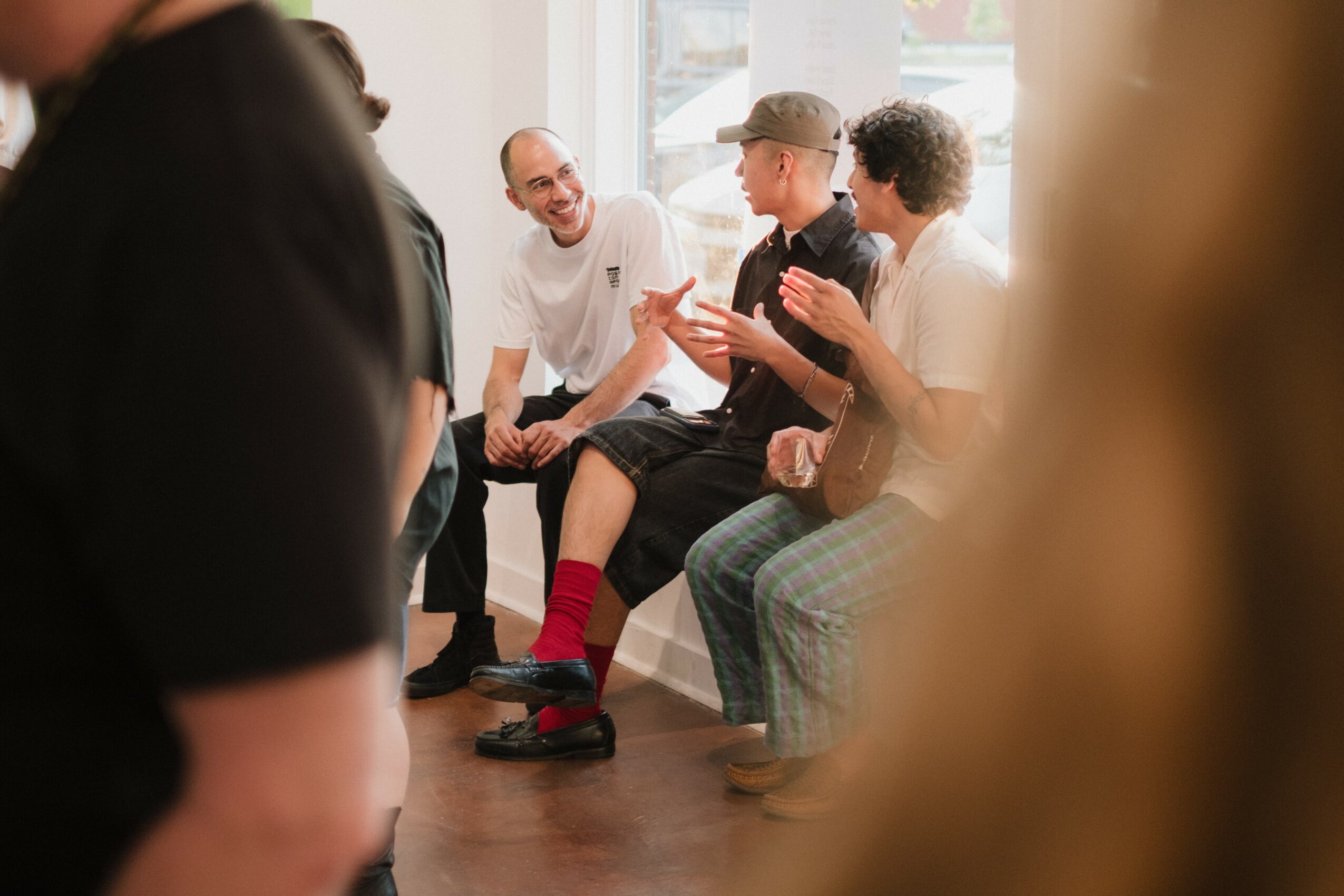 Group of people sitting and talking indoors, some smiling and gesturing, with sunlight coming through windows.