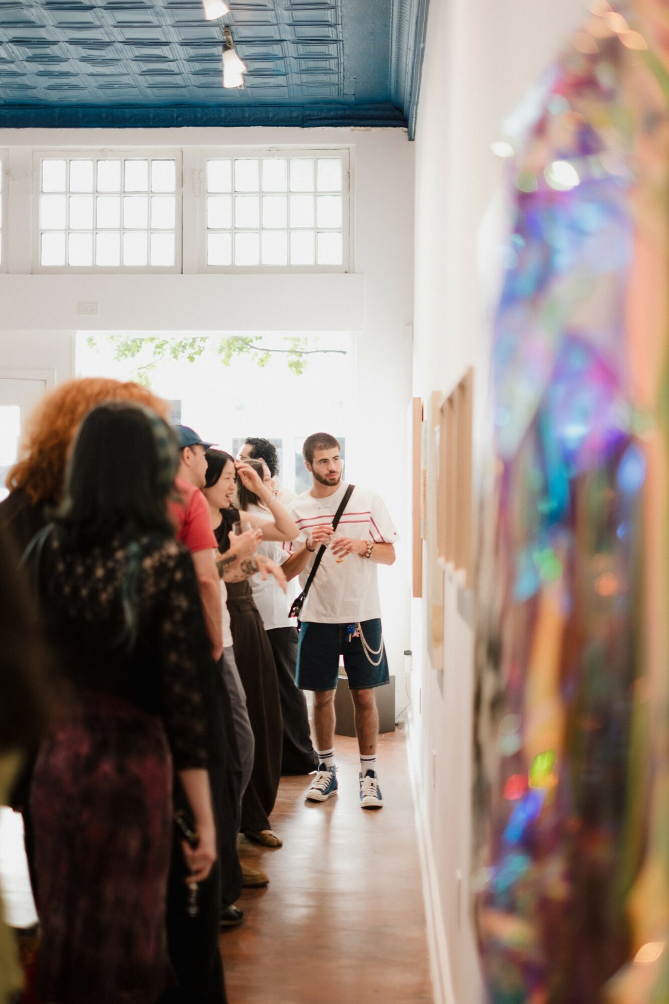 Group of people standing in a hallway, listening to a person playing a guitar, with a colorful display on the right side.