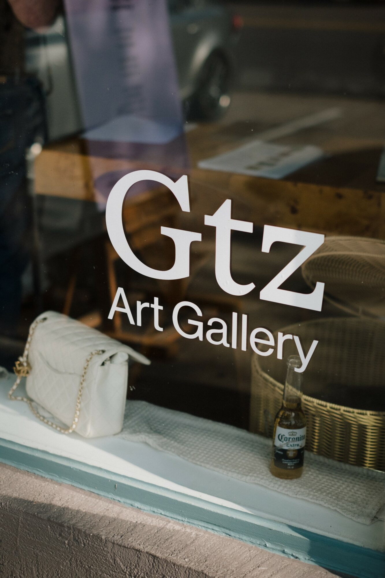 Gallery window with white text, a white purse, and a small bottle inside, reflecting street scene outside.