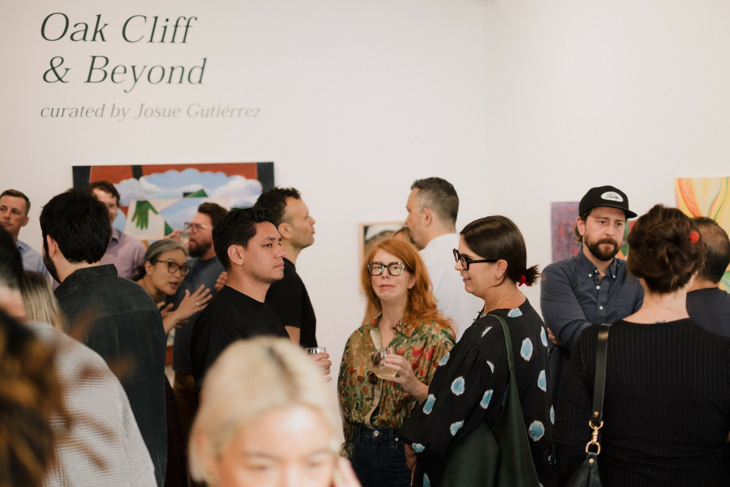 Group of people socializing at an art exhibition, with a white wall and artwork in the background.