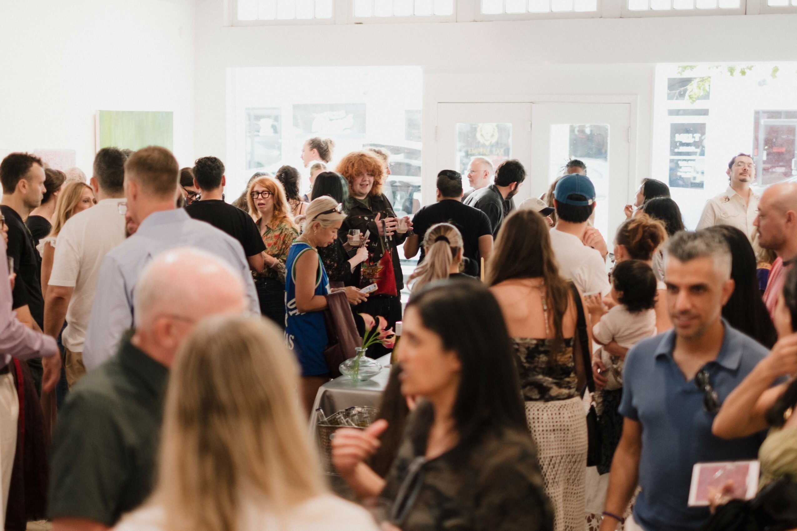 Crowd of people gathered indoors, some talking, some looking around, with large windows in the background.