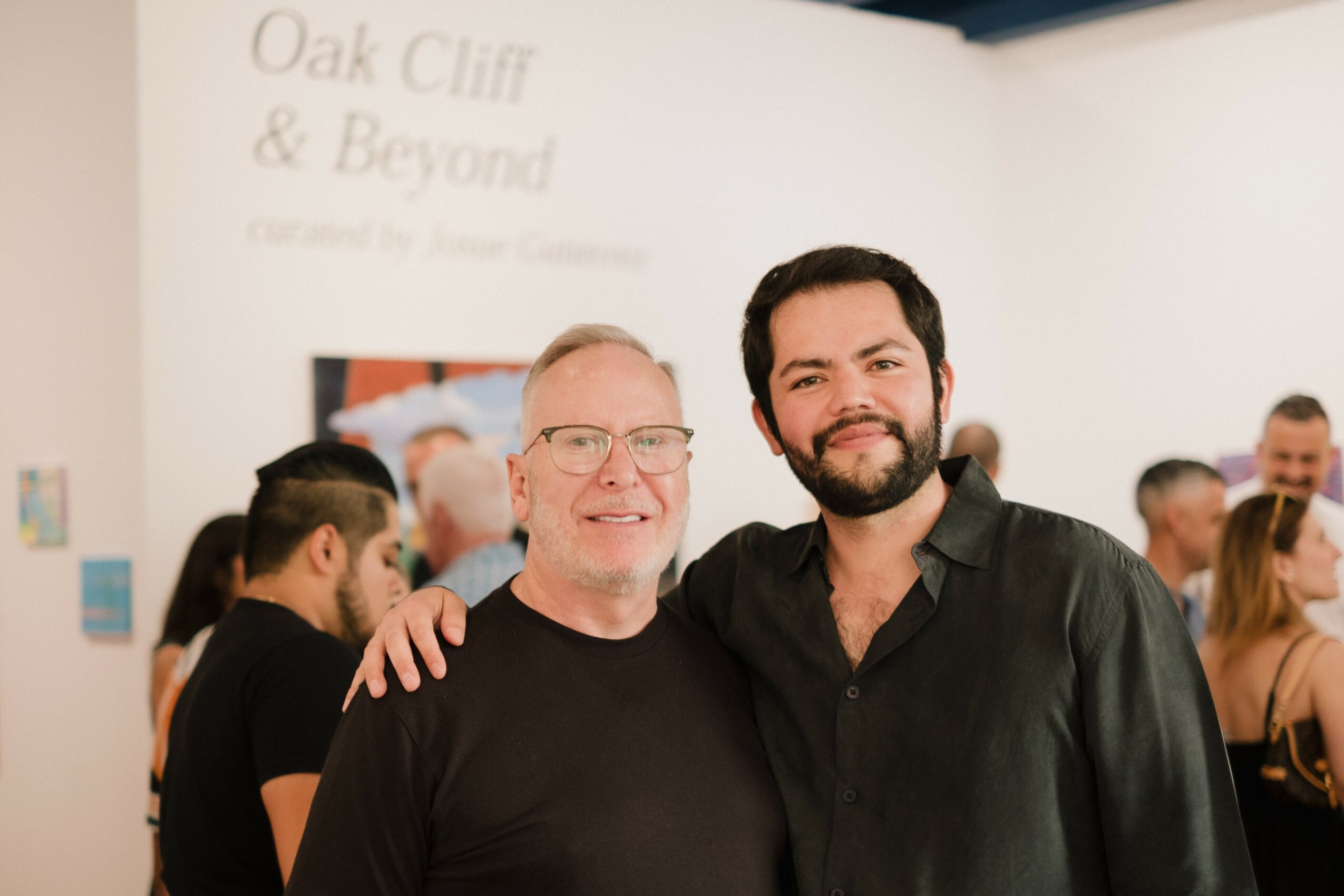 Two men smiling and standing close together in a crowded indoor event, with a sign reading 'Oak Cliff & Beyond' in the background.