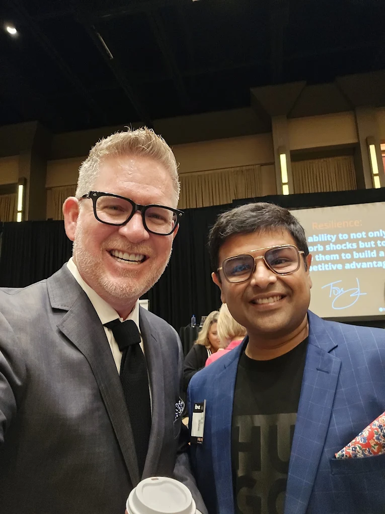 Two men in suits and glasses smiling at the camera, standing indoors with a dark ceiling and a screen in the background.