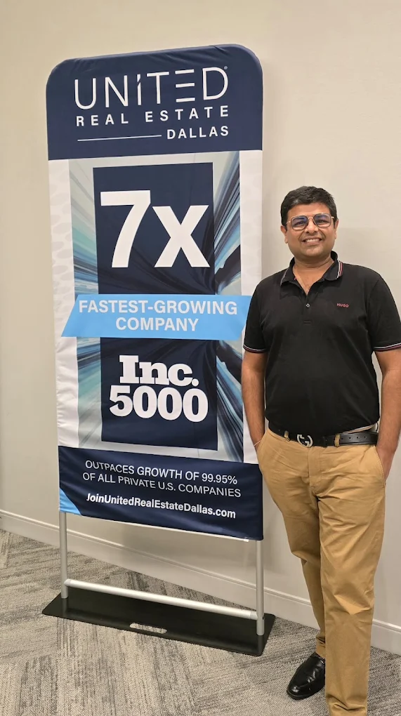 Man standing next to a tall banner with text about a real estate company, smiling, wearing a black polo and khakis.