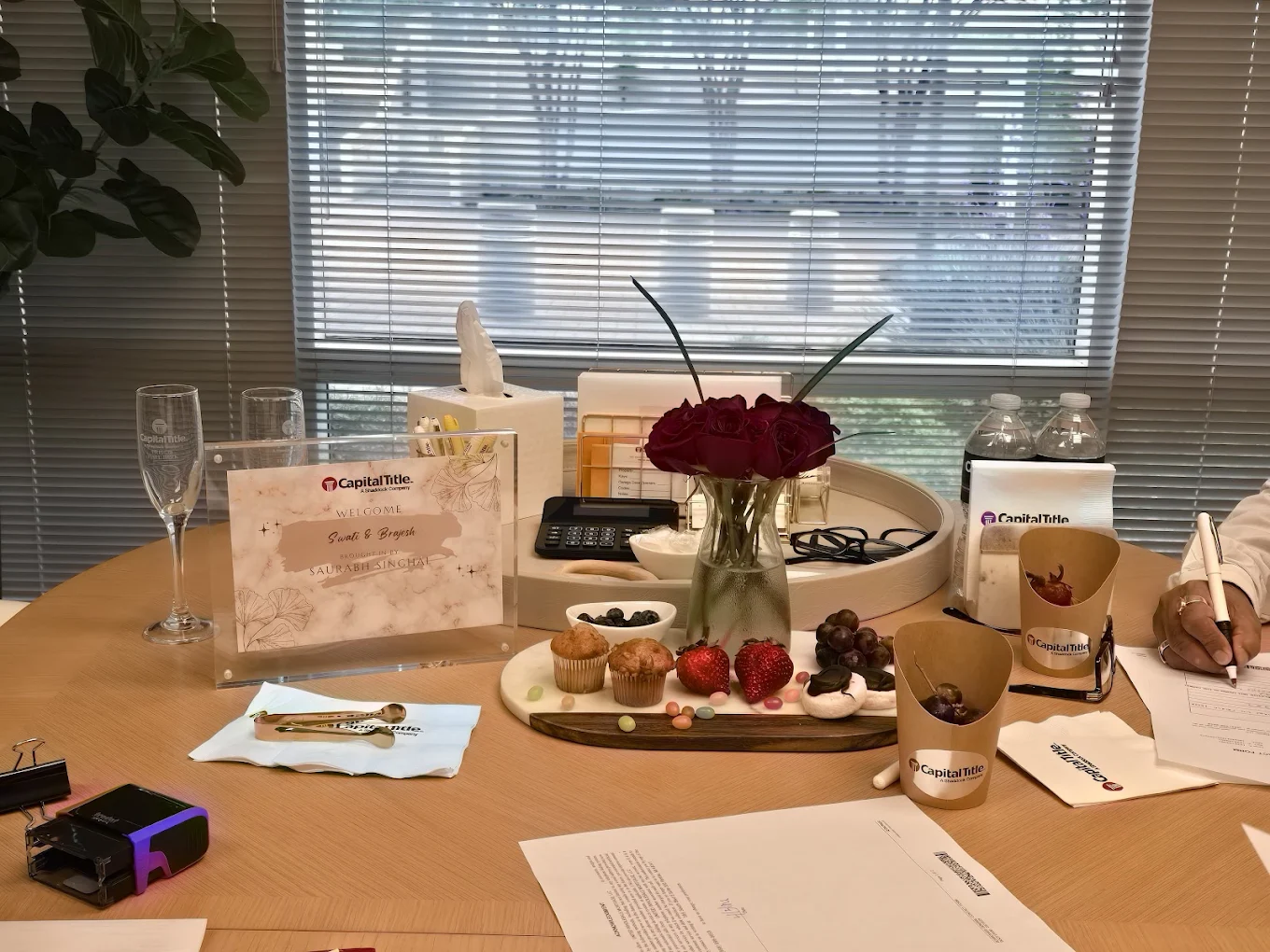 Table with a flower vase, glasses, cups, and papers in front of a window with blinds.