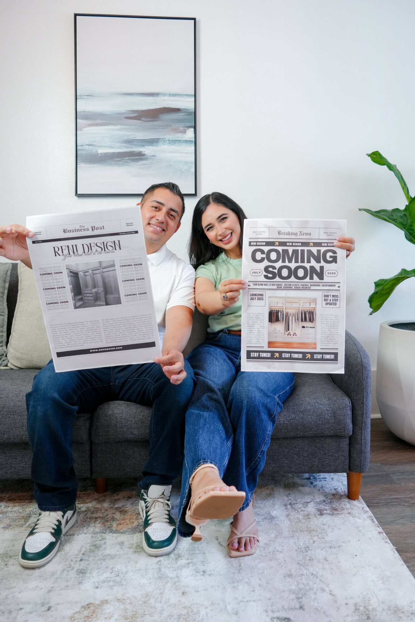 Two people sitting on a sofa holding newspapers, smiling, in a bright living room with a painting and plant.