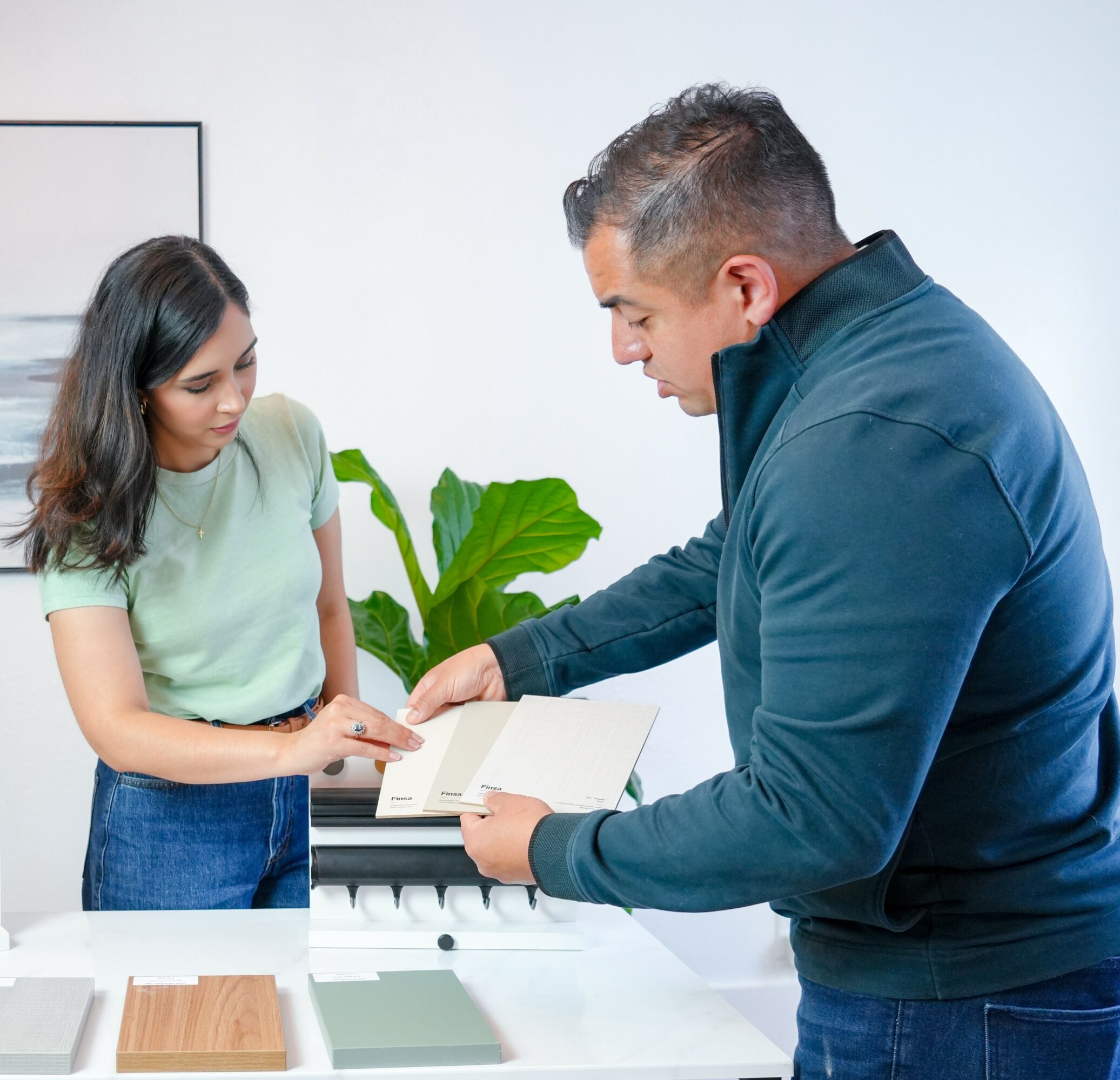 Two people exchanging a book in a bright room with a plant and framed picture, one person handing the book to the other.
