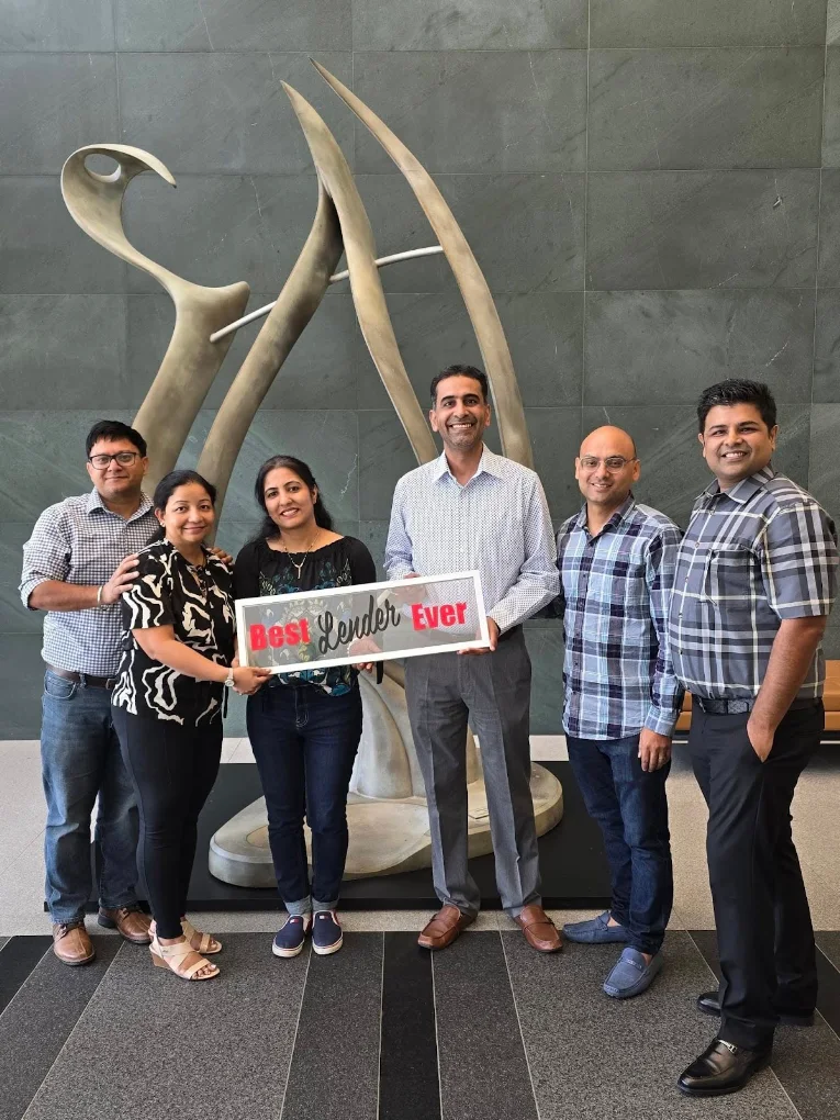 Group of six people standing in front of a large abstract sculpture, holding a sign that reads 'Best' and 'Even'.