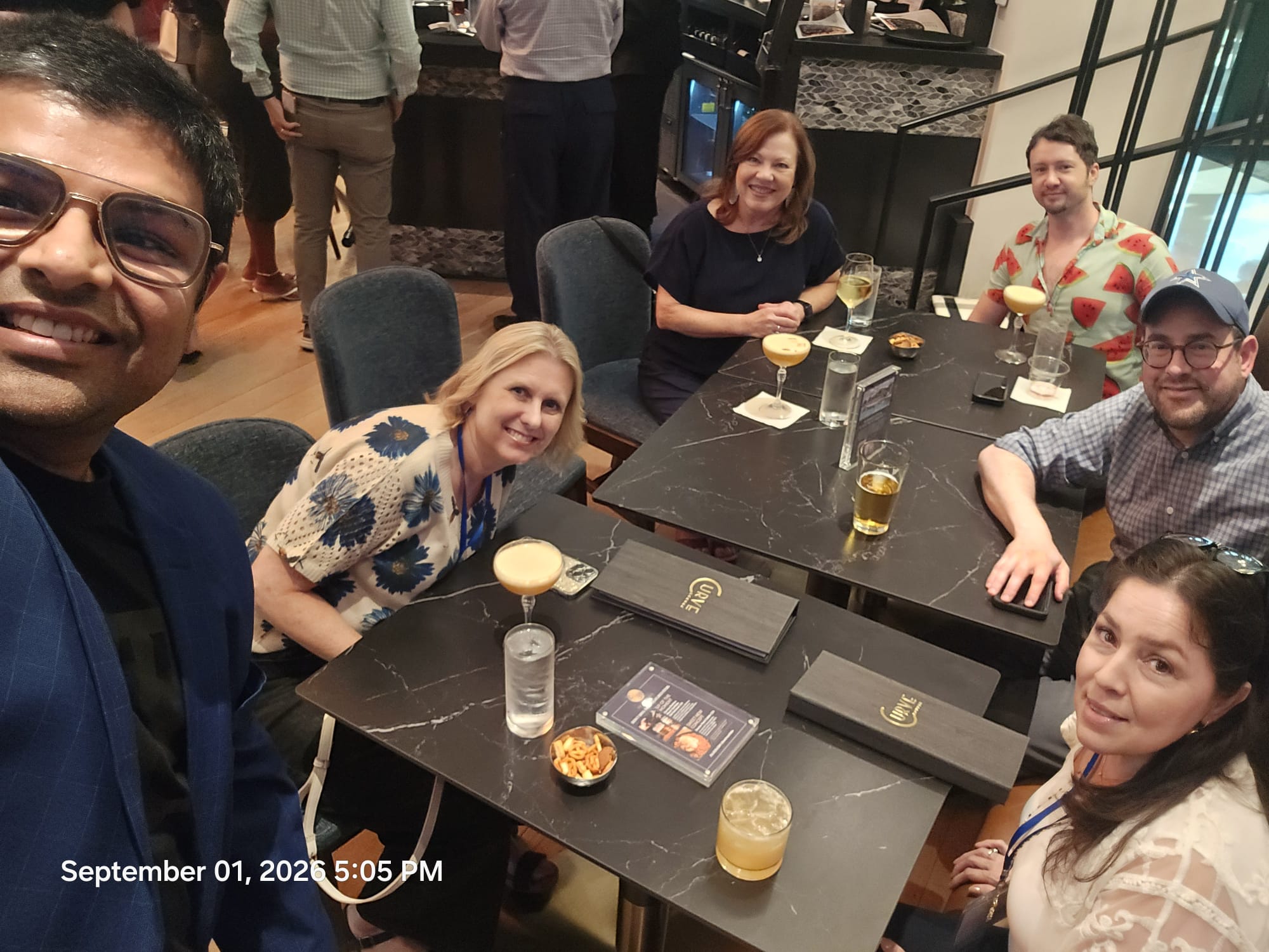 Group of five people sitting at a table in a restaurant, smiling and enjoying drinks and food.