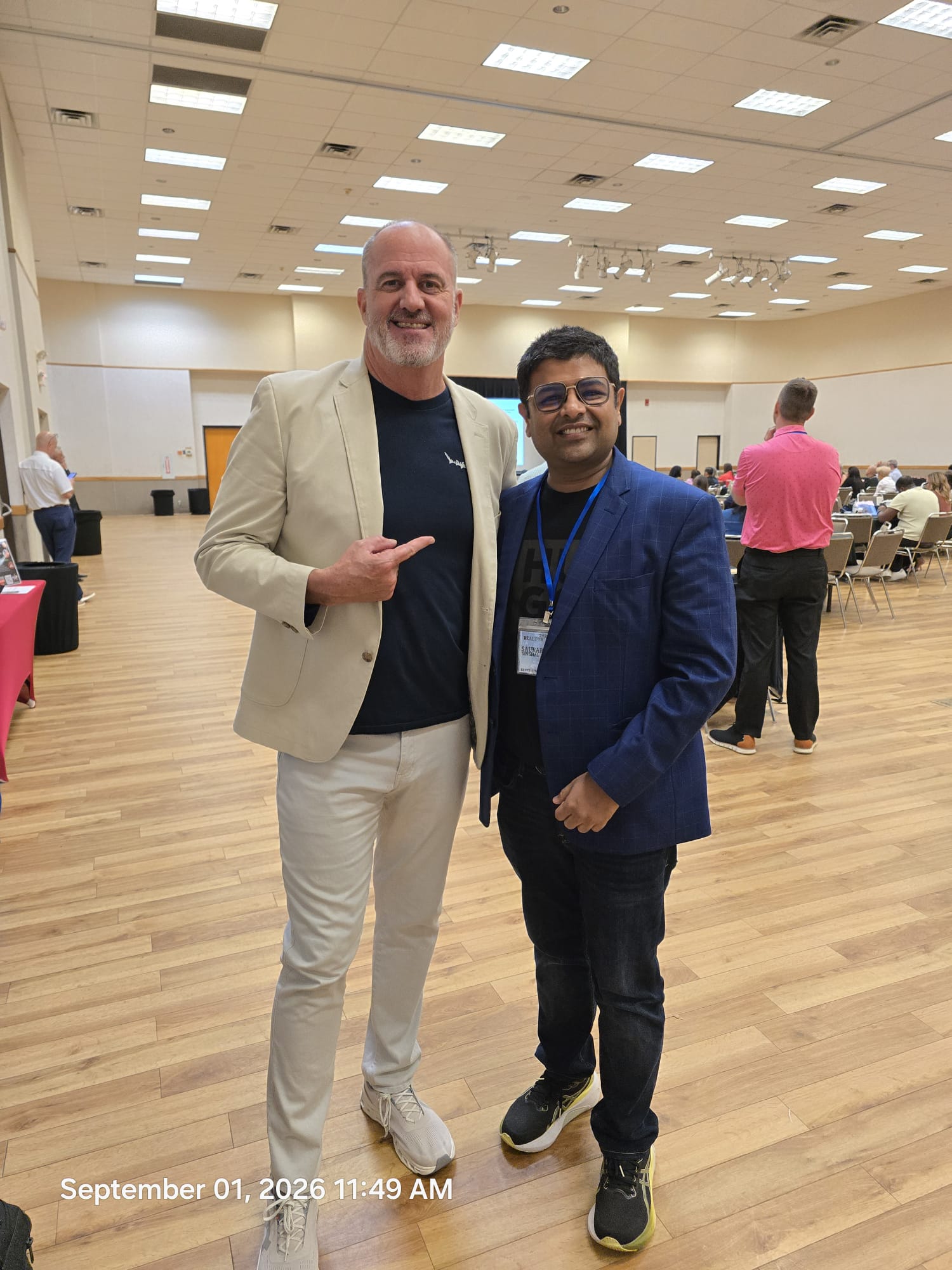 Two men standing together in a large room, smiling, with other people in the background, wooden floor, ceiling lights, and a date and time stamp.