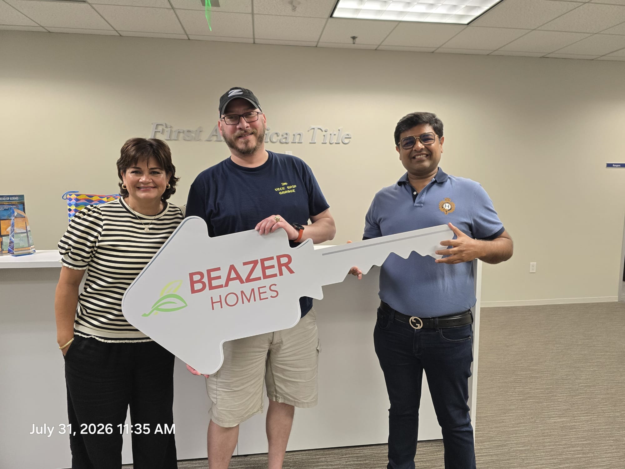 Three people standing indoors, holding a large key-shaped sign that reads 'BEAZER HOMES'.