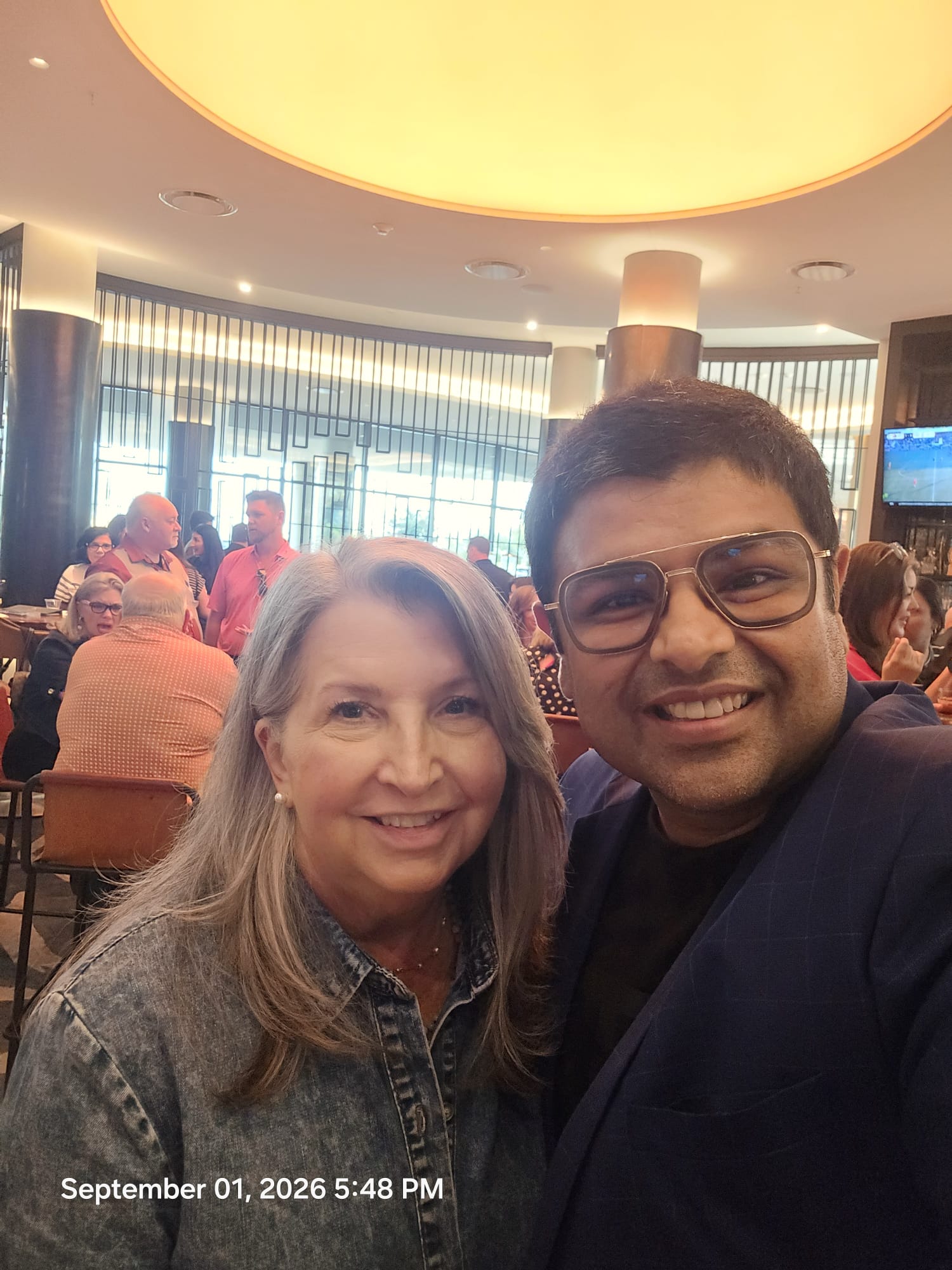 Two people smiling in a restaurant with a large circular ceiling light and other diners in the background.