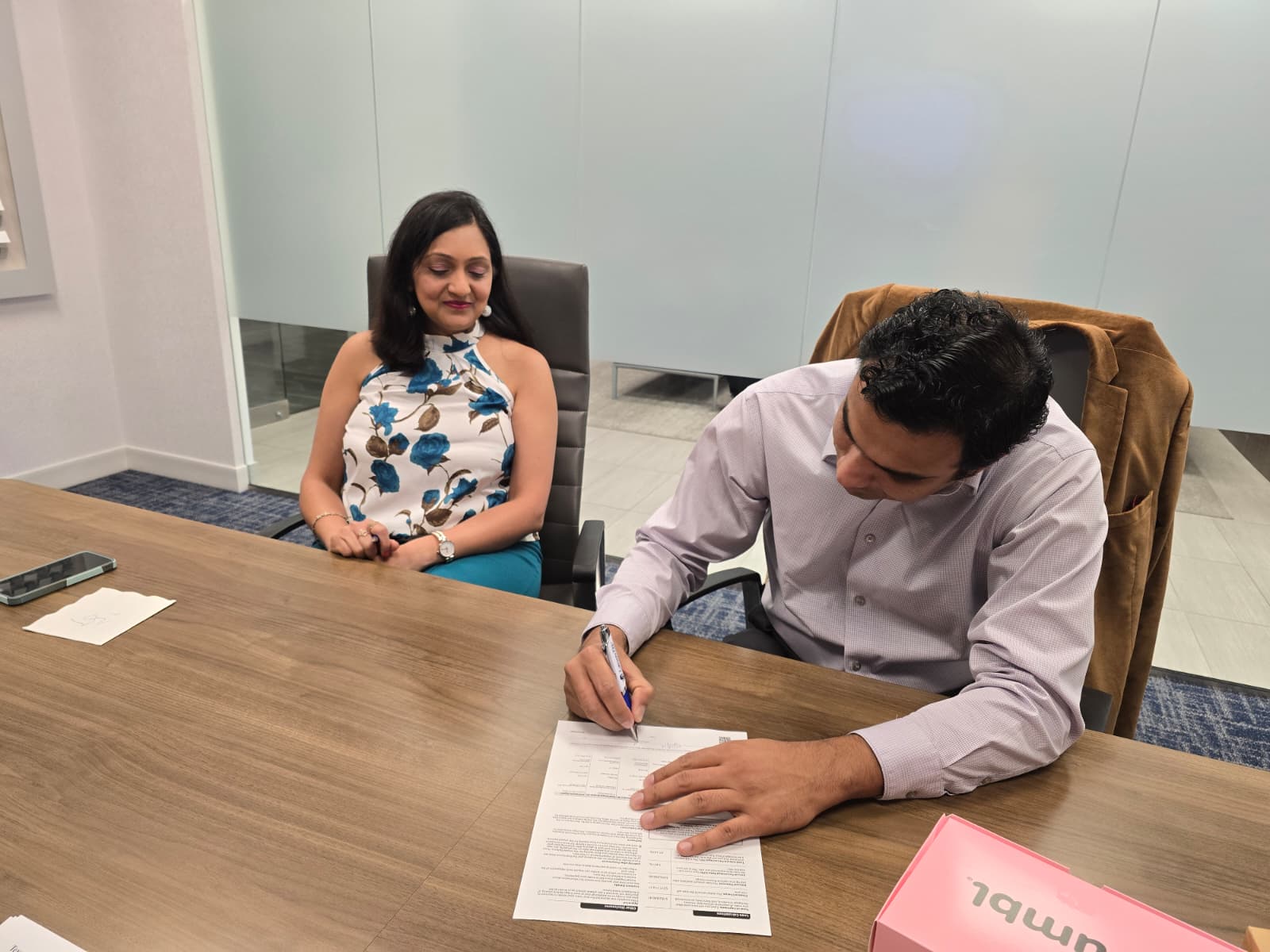 A woman sitting at a conference table with a man writing on a paper, in a modern office setting.