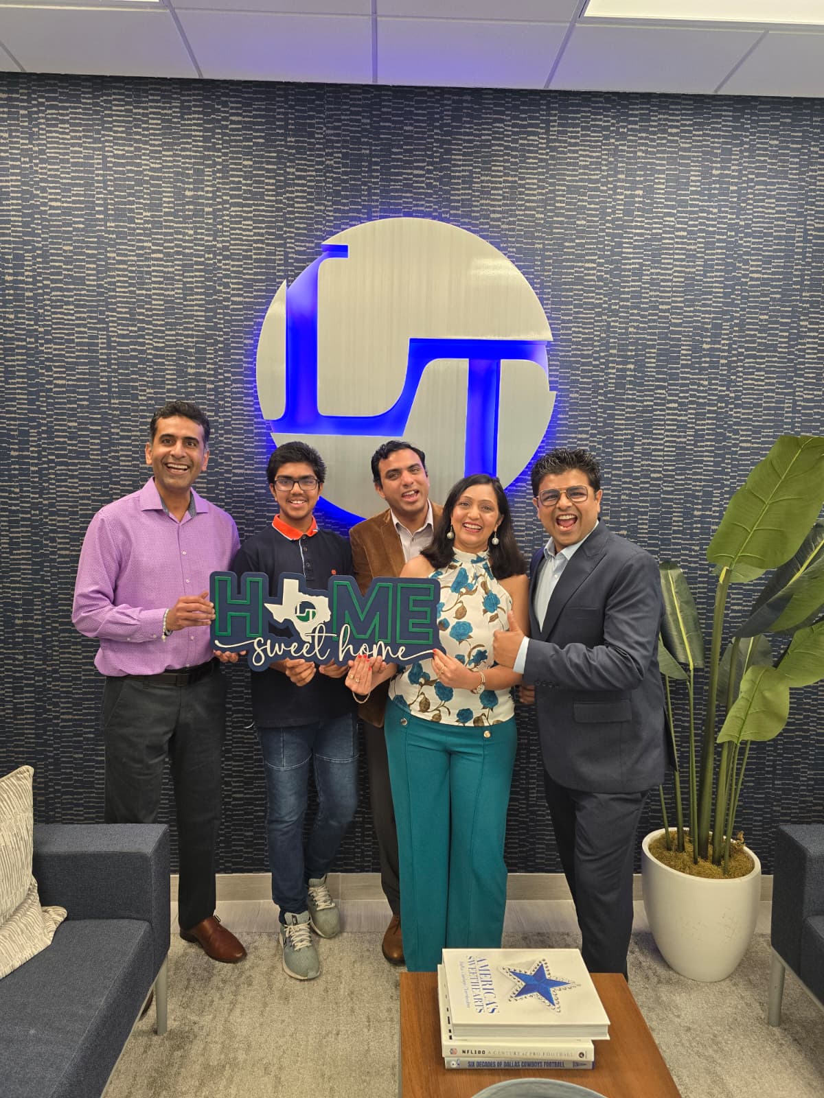 Five people standing in front of a wall with a large logo, holding a sign that says 'HIS HOME Sweet Home,' with a potted plant nearby.