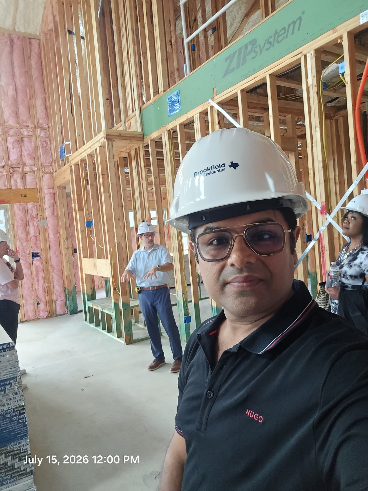 Person wearing safety helmet and glasses taking selfie at construction site with wooden framing in background.