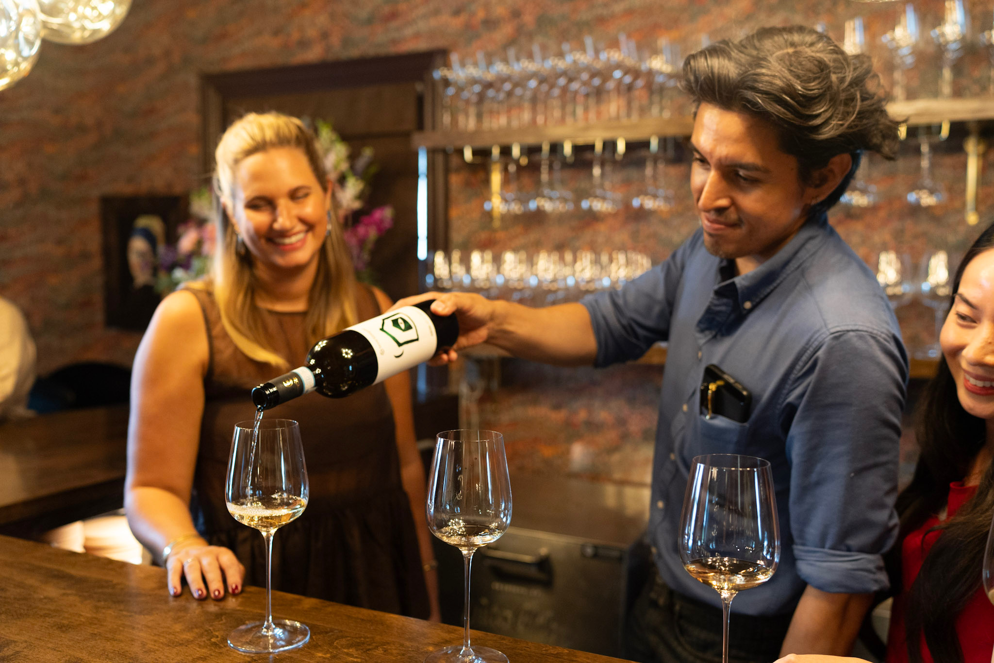 Bartender pours wine for smiling woman at bar with glasses and brick wall background.