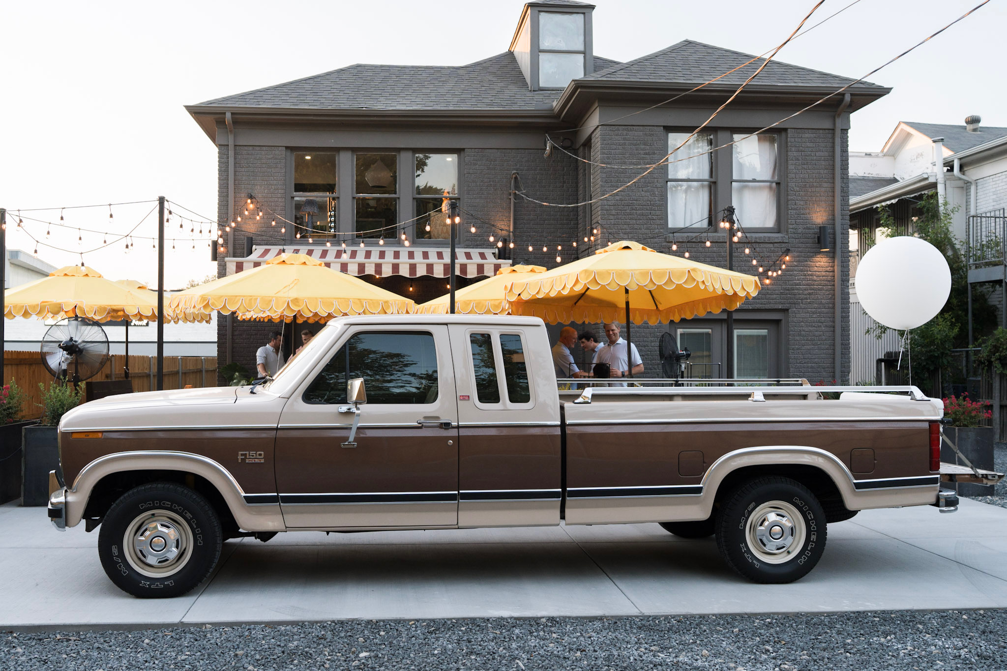 Brown and white pickup truck parked in front of a dark gray house with string lights and yellow umbrellas.