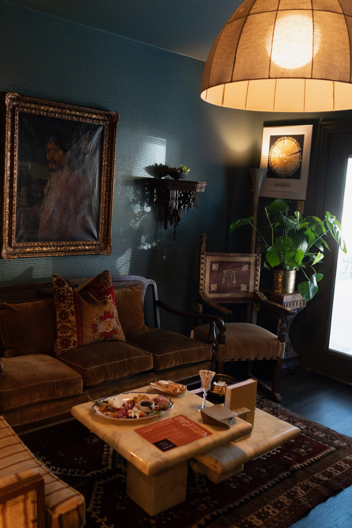 Living room with a brown sofa, framed painting, potted plant, and a marble coffee table with snacks and books, illuminated by a large hanging lamp.