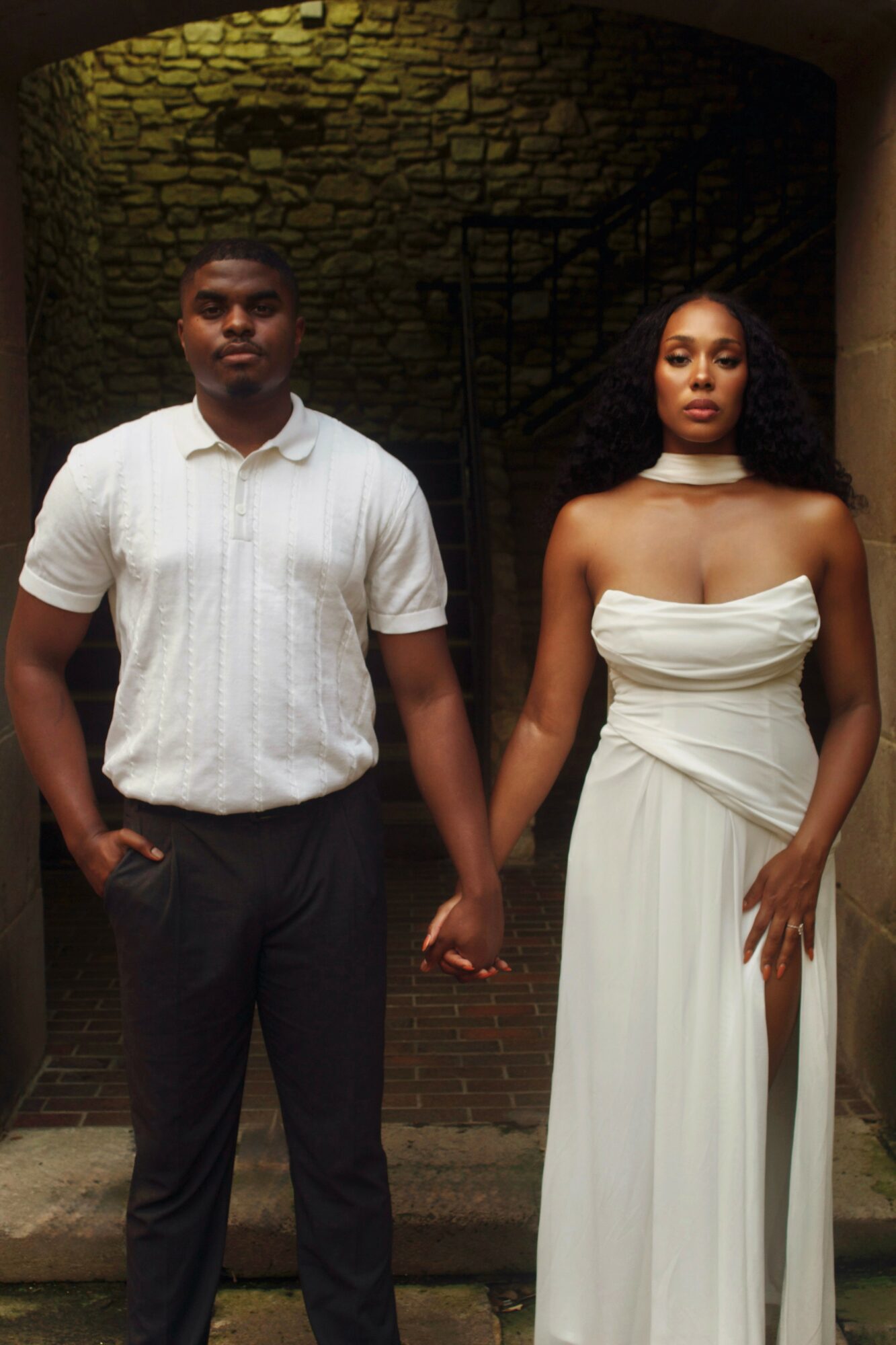 A man and woman holding hands, standing outdoors in front of a brick wall and stone archway, dressed formally.