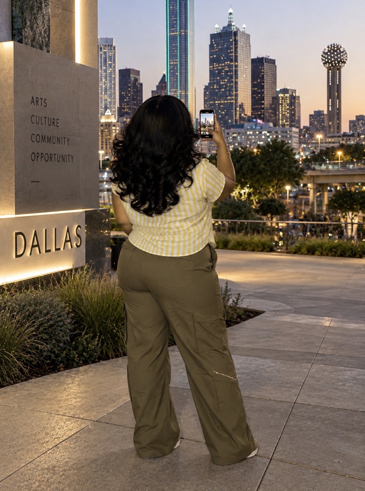 Person with long dark hair taking a photo of the city skyline at dusk, standing on a paved area with trees and a sign that reads 'DALLAS'.