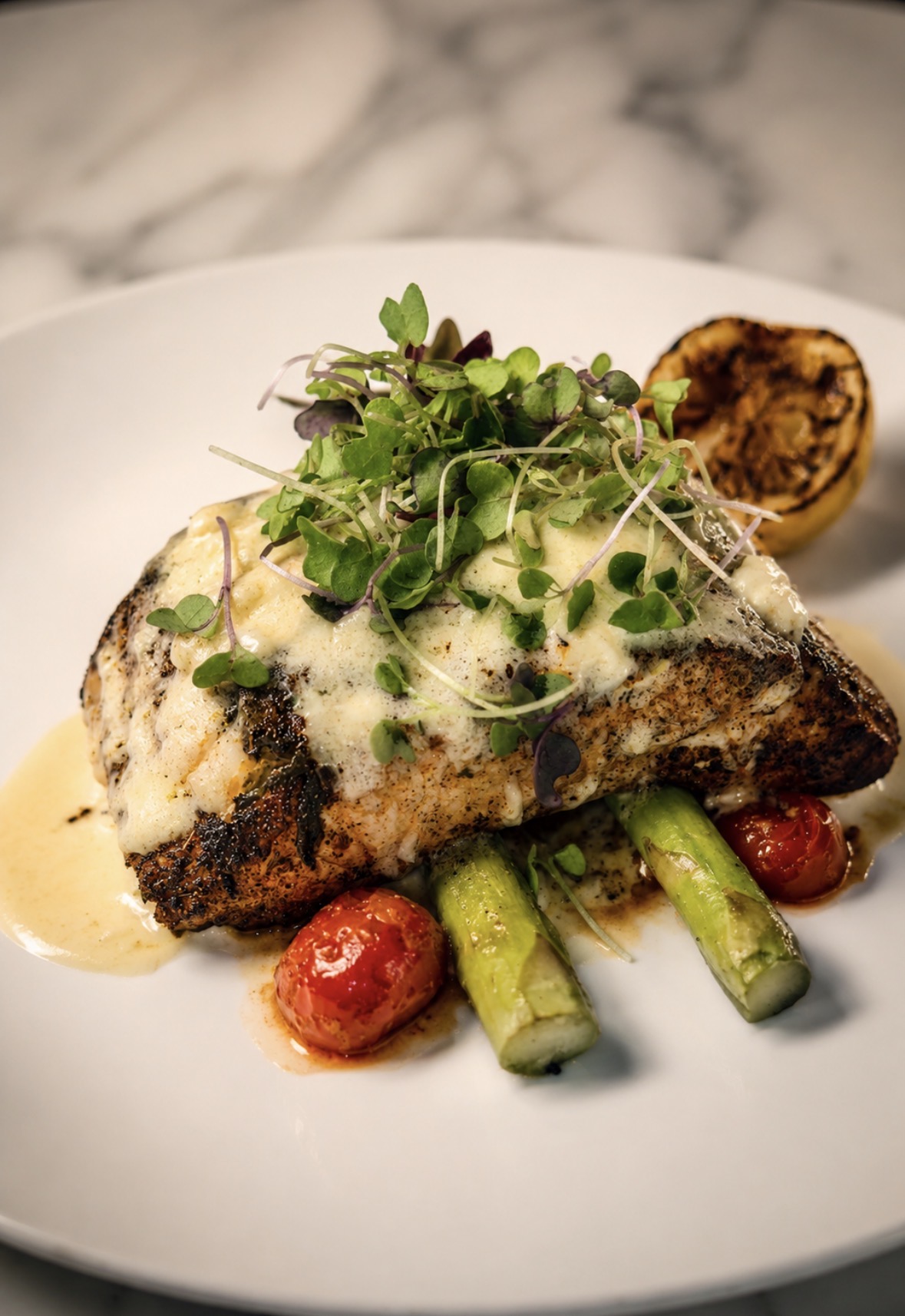 Grilled fish fillet topped with microgreens, served with cherry tomatoes, asparagus, and a lemon half on a white plate.