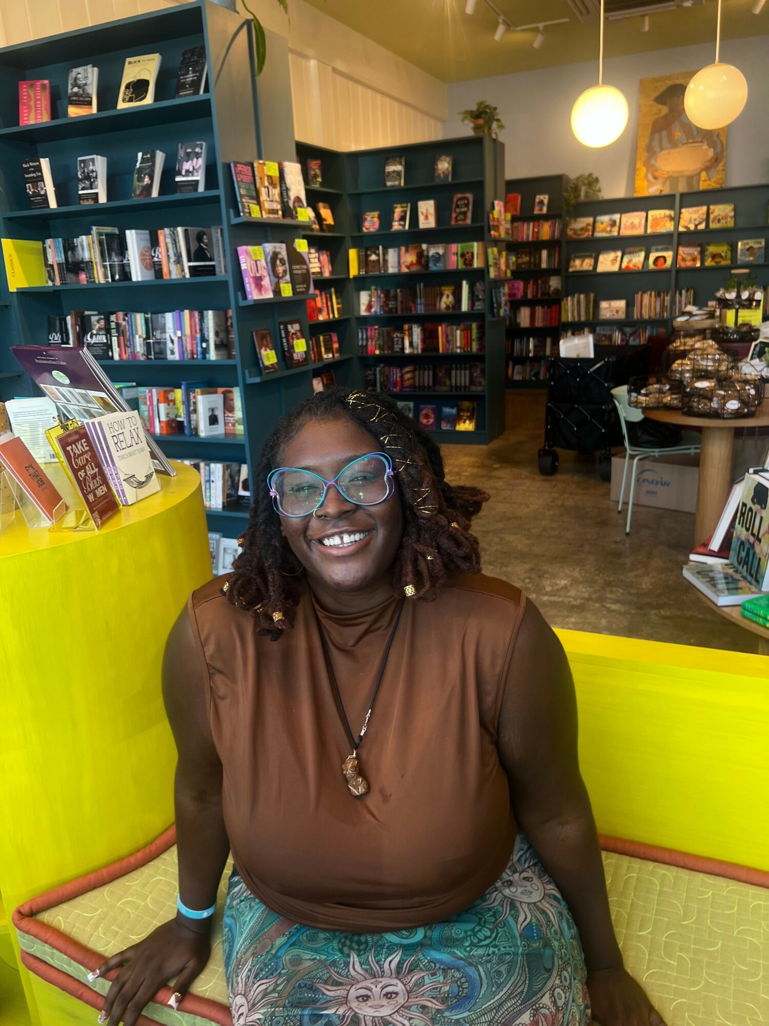 Woman with glasses smiling, sitting on a yellow bench in a bookstore, surrounded by shelves of books.