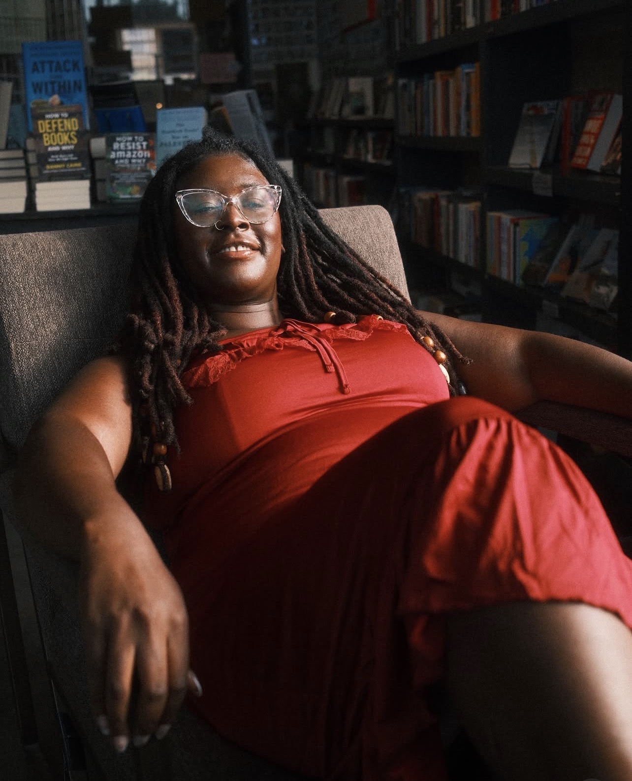 Woman with glasses in a red dress relaxing in a library, smiling, surrounded by bookshelves.