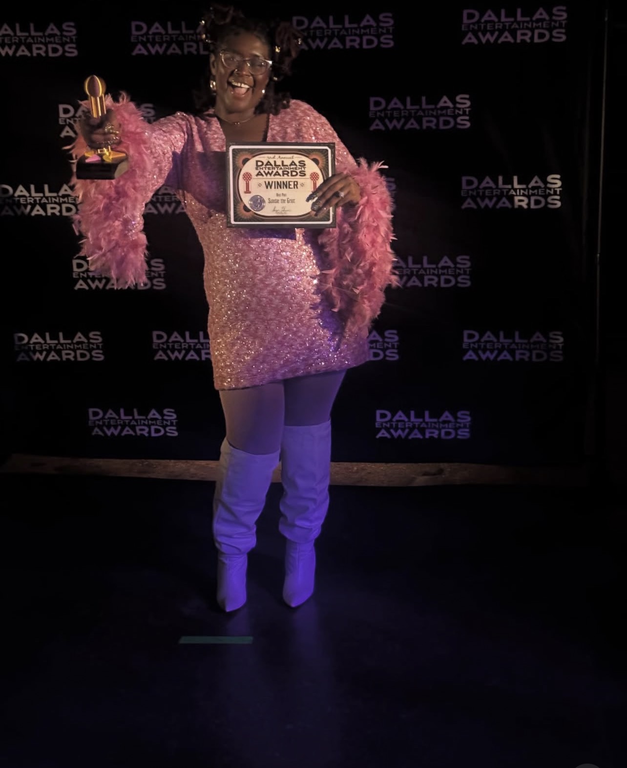 Person holding an award and certificate, smiling, wearing a pink dress with feathered sleeves, standing in front of a Dallas Awards backdrop.