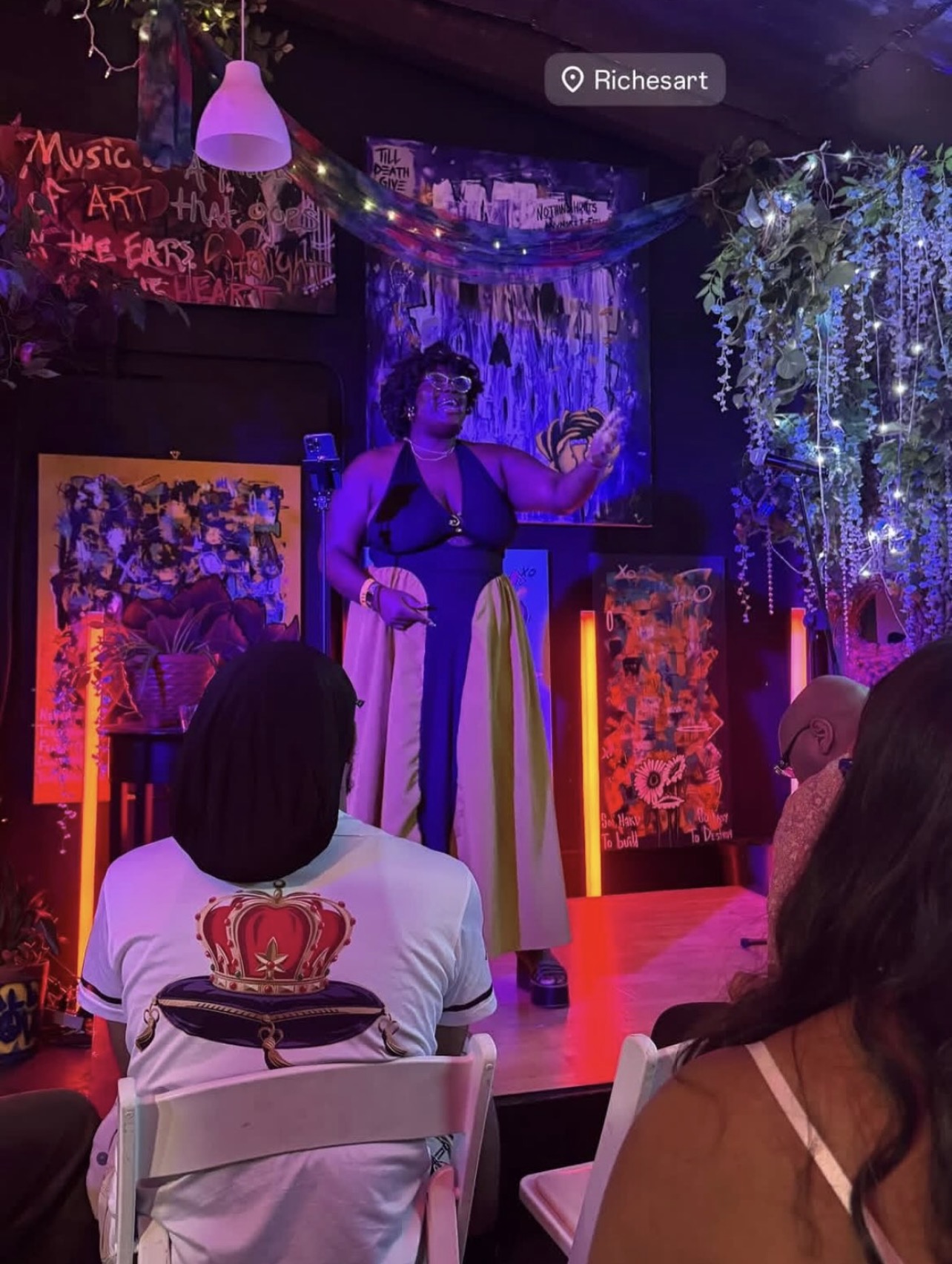 Performer on stage with microphone, audience seated, colorful background with hanging lights and artwork, indoor setting.
