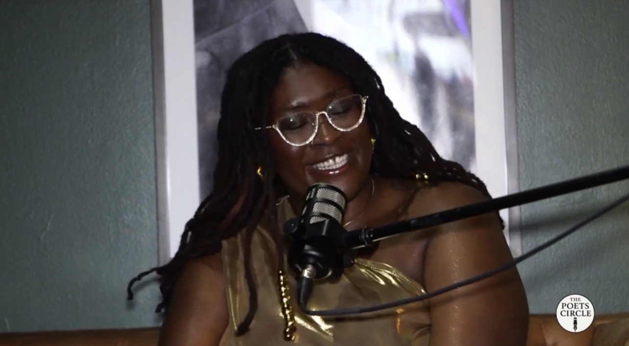 Woman with glasses smiling, speaking into a microphone, in a room with a framed picture behind her.