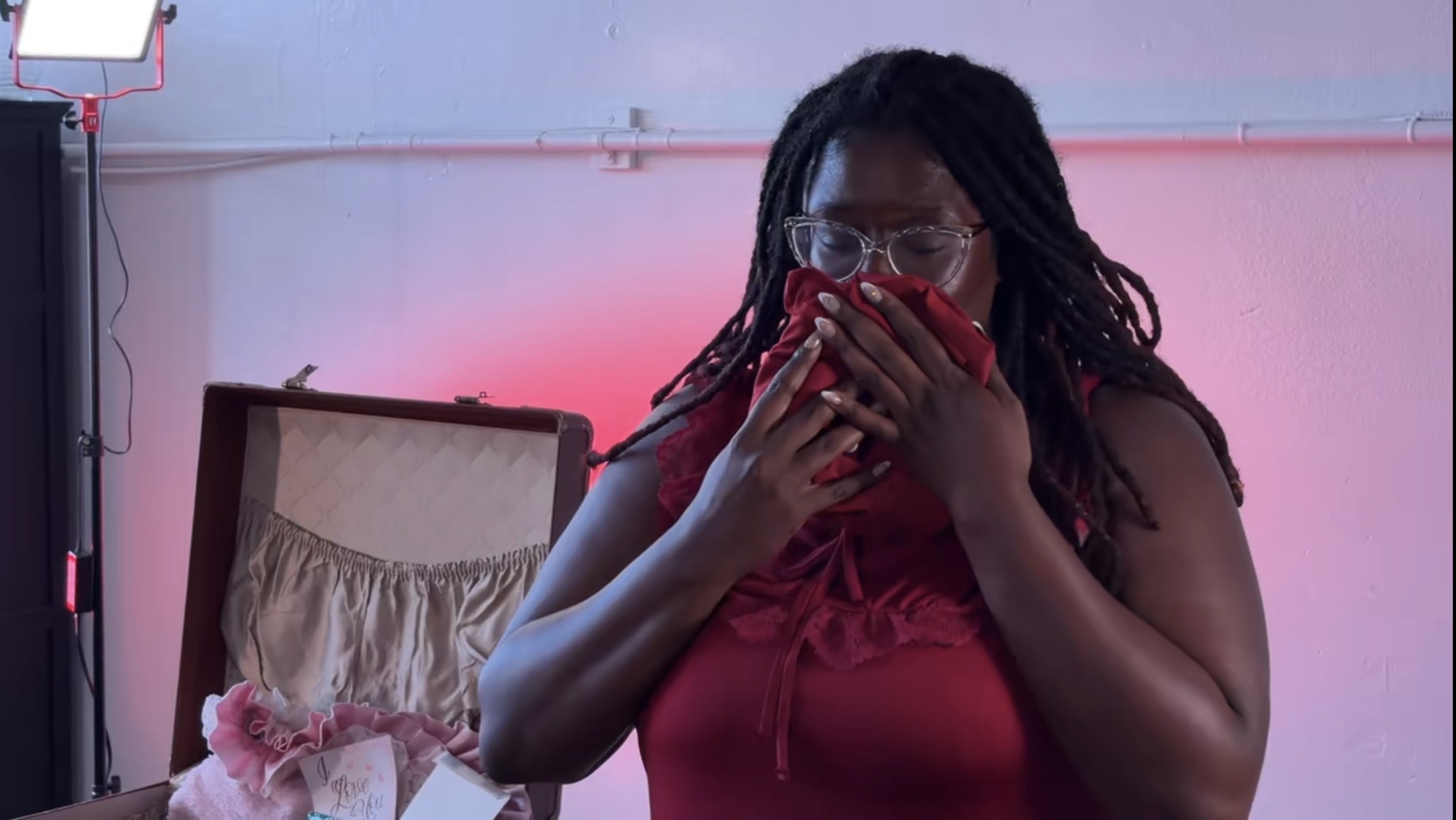 Woman with glasses and dreadlocks covering her face with hands, wearing a red top, in a room with pink lighting.