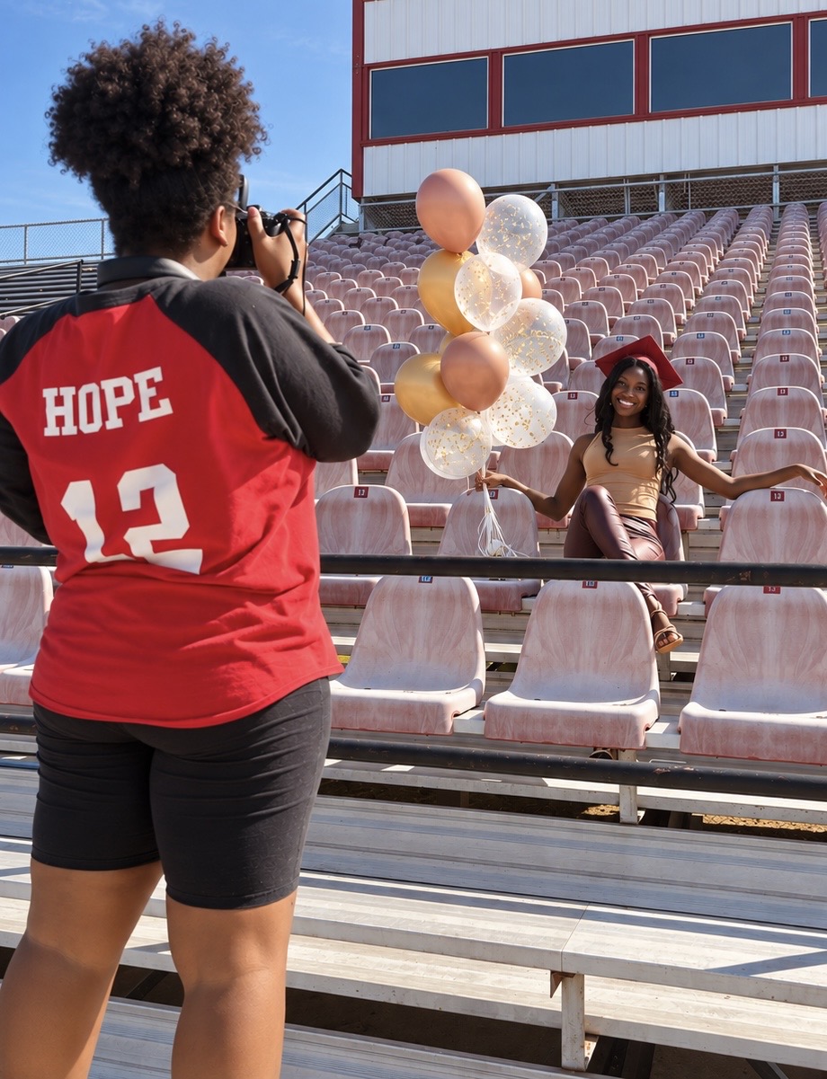 Person with curly hair taking a photo of a girl holding balloons in an outdoor stadium seating area.
