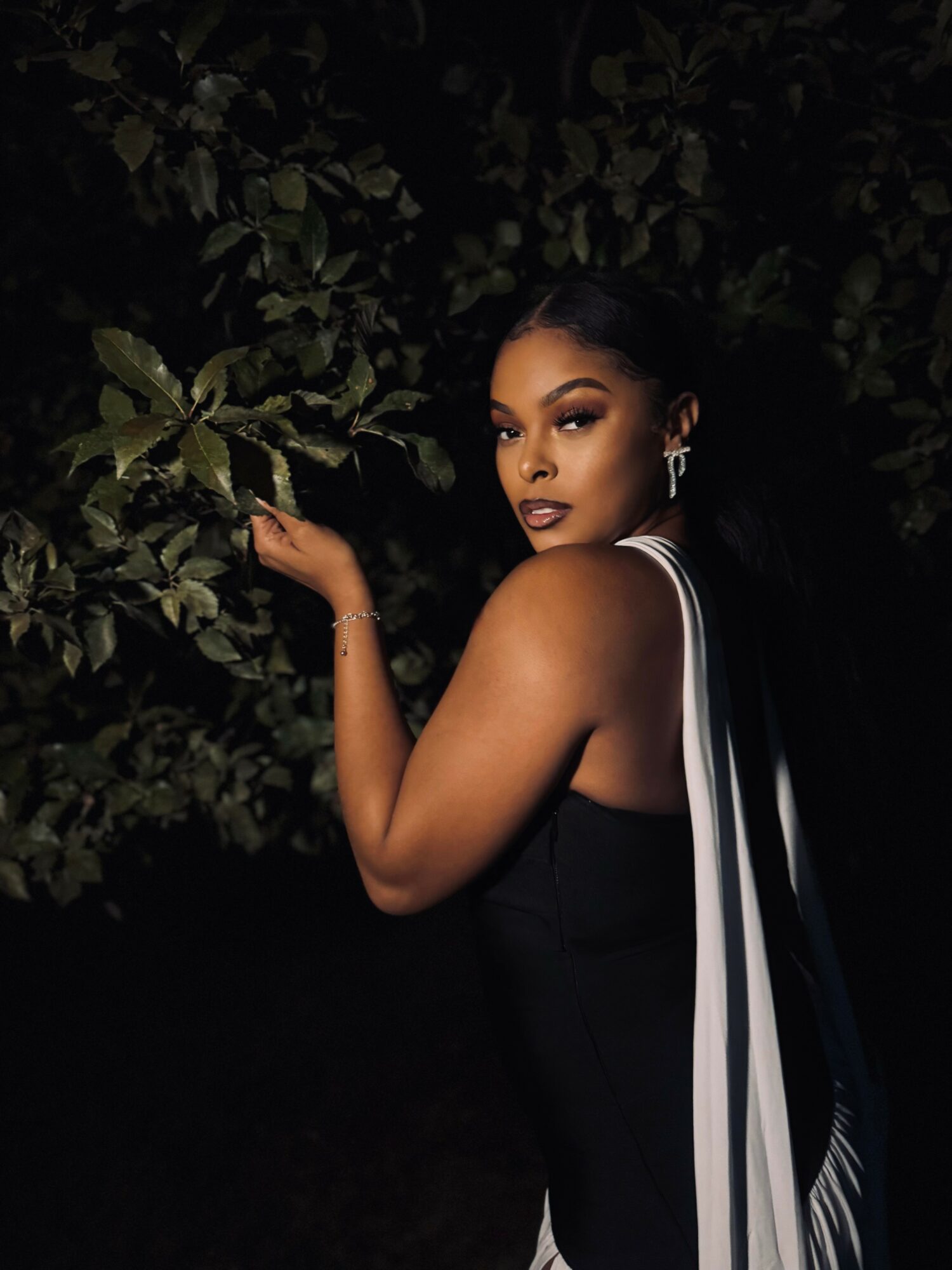 Woman with dark hair and earrings poses outdoors at night, surrounded by leaves, wearing a black dress with white accents.