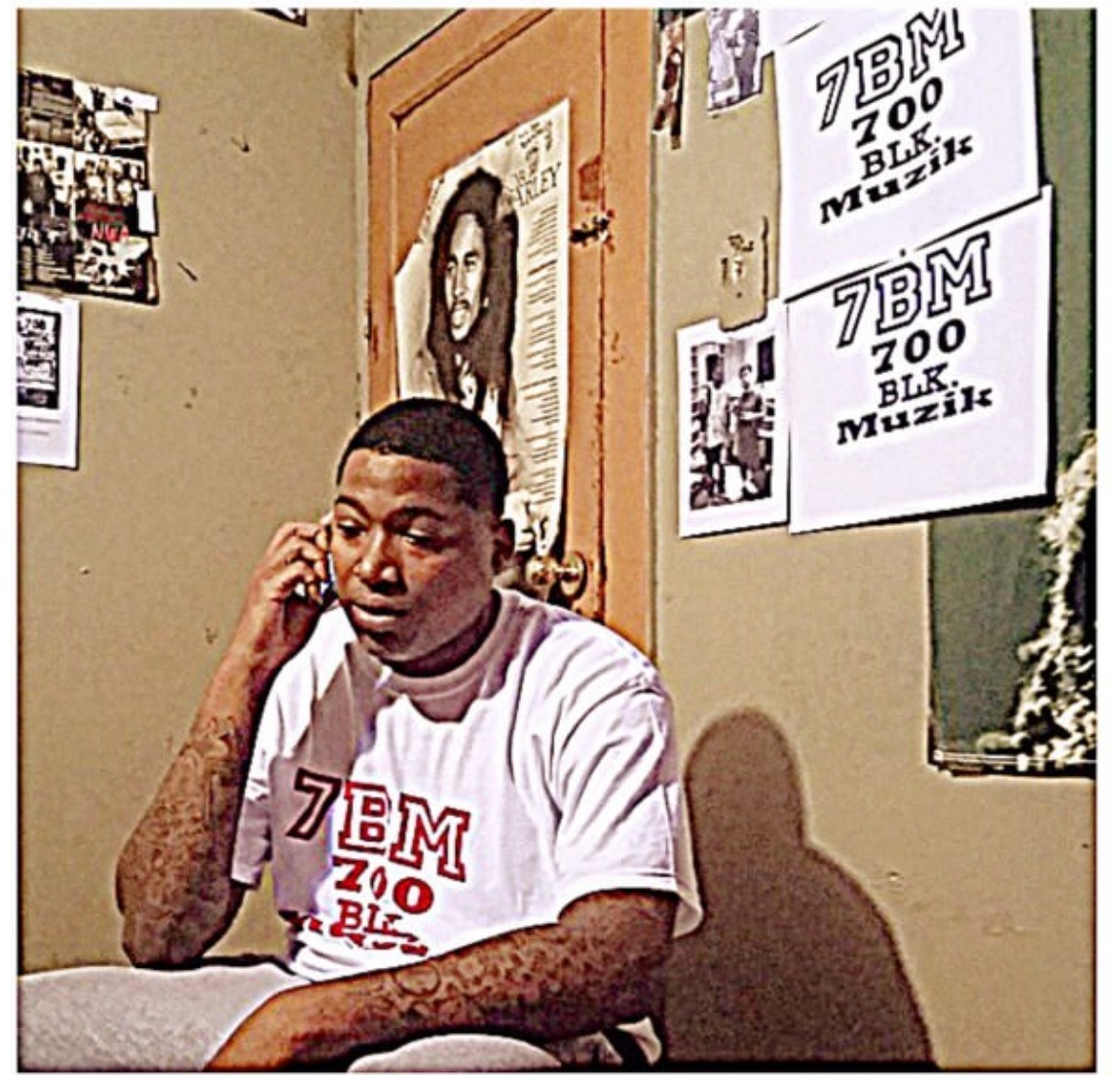 Young man sitting at a desk, talking on a phone, wearing a white T-shirt with red and black text, in a room with posters and a door.