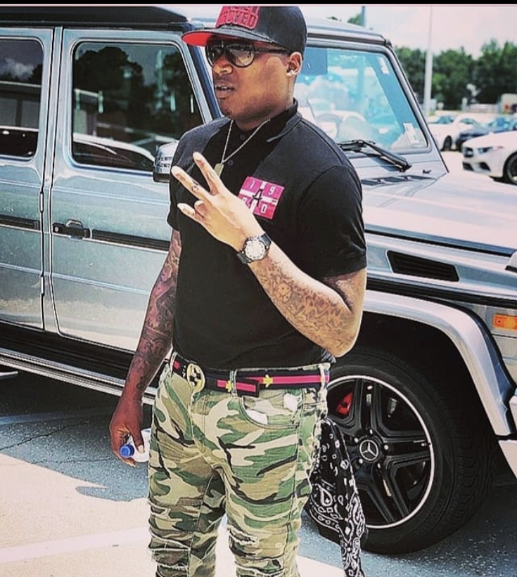 Man with tattoos wearing glasses, a black cap, black shirt, camouflage pants, and a watch, standing next to a black vehicle.