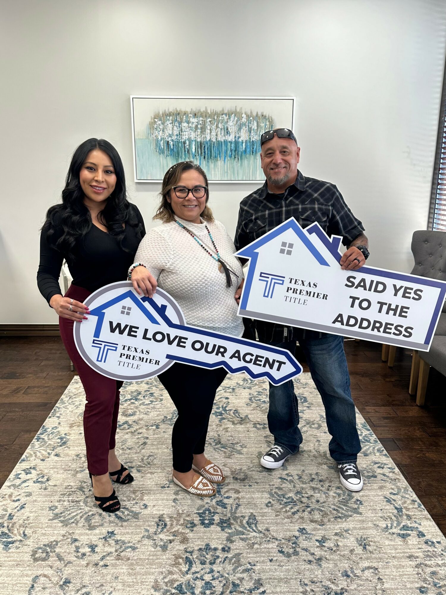 Three people standing indoors holding signs, smiling, with a white wall and window behind them.