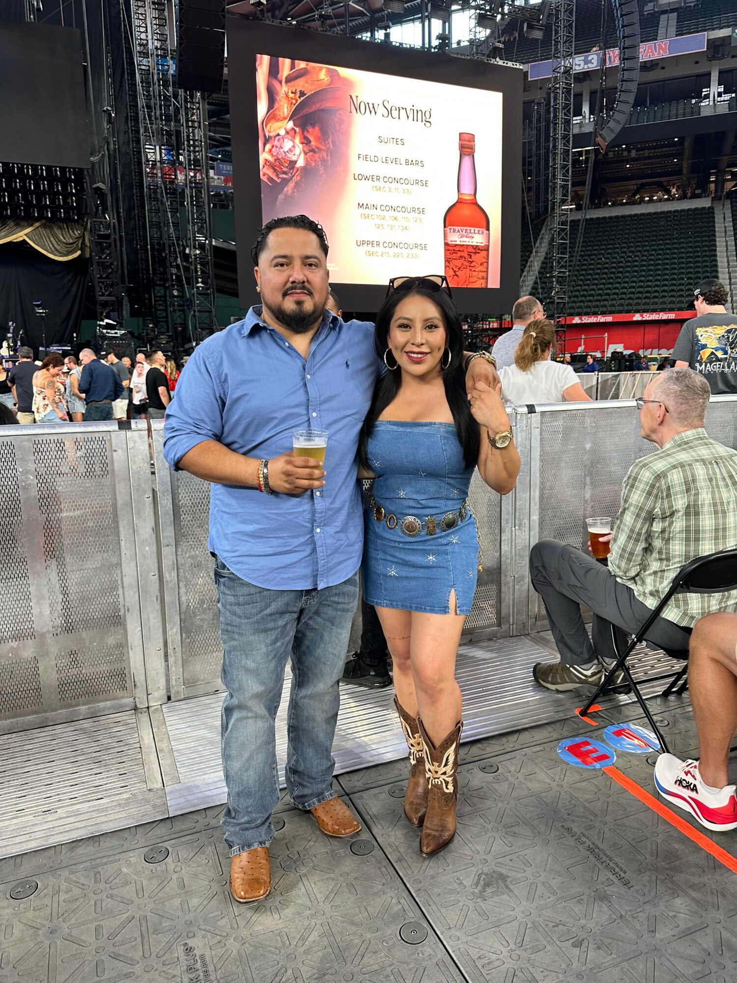A man and woman stand together at an outdoor event, holding drinks, with a stage and large screen in the background.