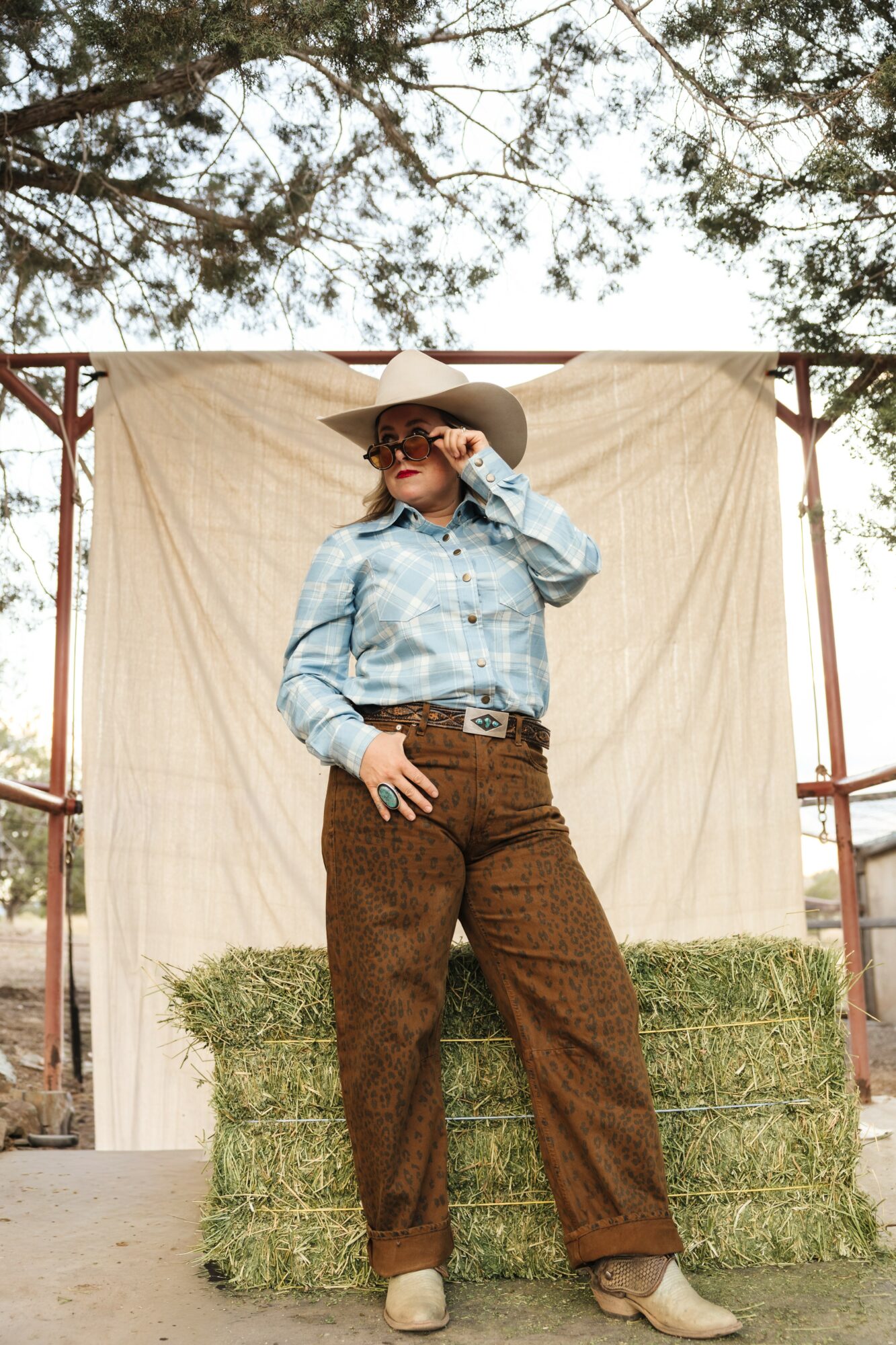 Person wearing a wide-brimmed hat, sunglasses, denim shirt, and brown pants, standing outdoors with hay bales and a backdrop.