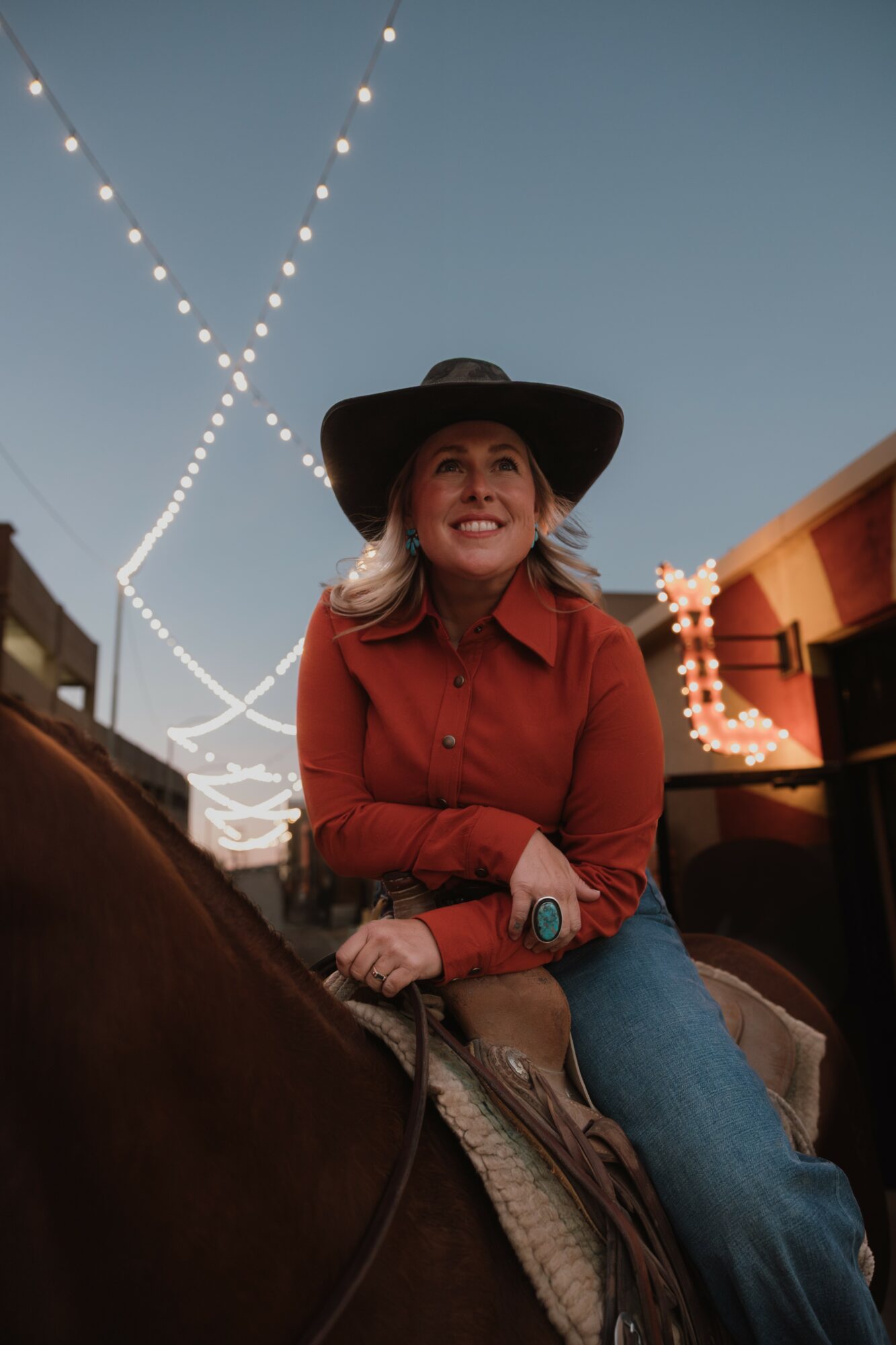 Woman wearing a wide-brimmed hat and red shirt, smiling outdoors at dusk with string lights overhead.