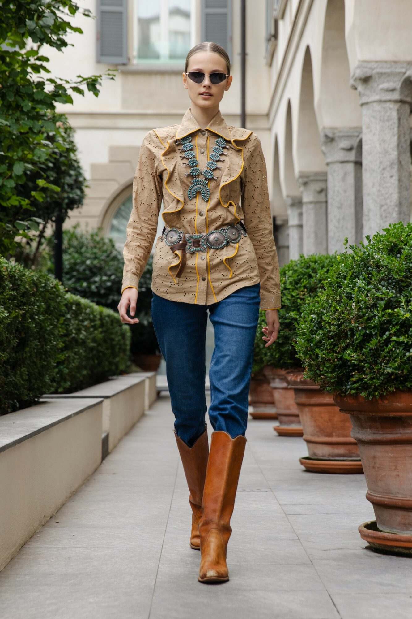 Woman walking outdoors in a city, wearing sunglasses, a patterned blouse, blue jeans, and brown boots, with greenery and buildings in background.