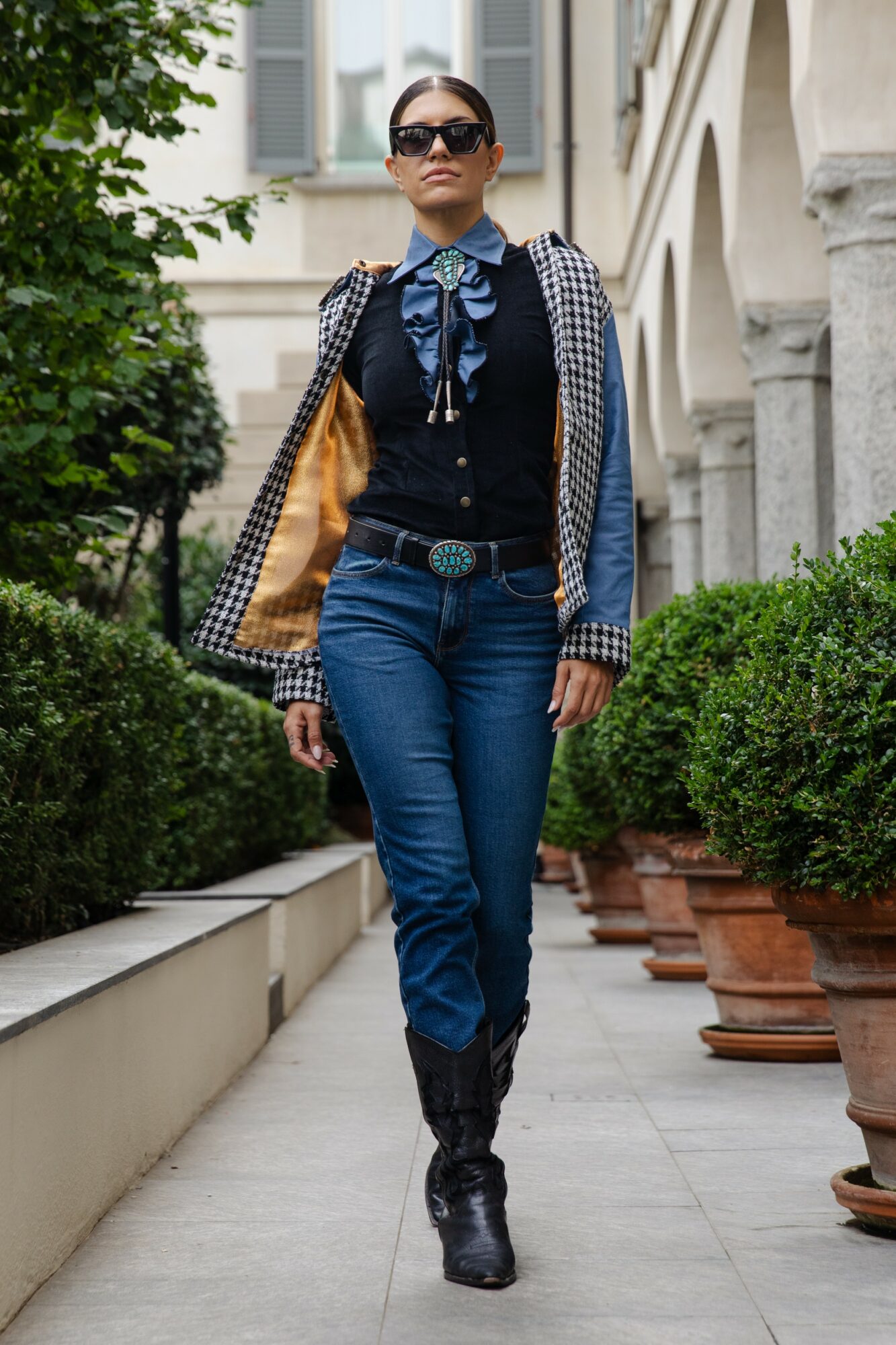 Woman walking outdoors in sunglasses, wearing a patterned jacket, blue jeans, and black boots, with potted plants and buildings in background.