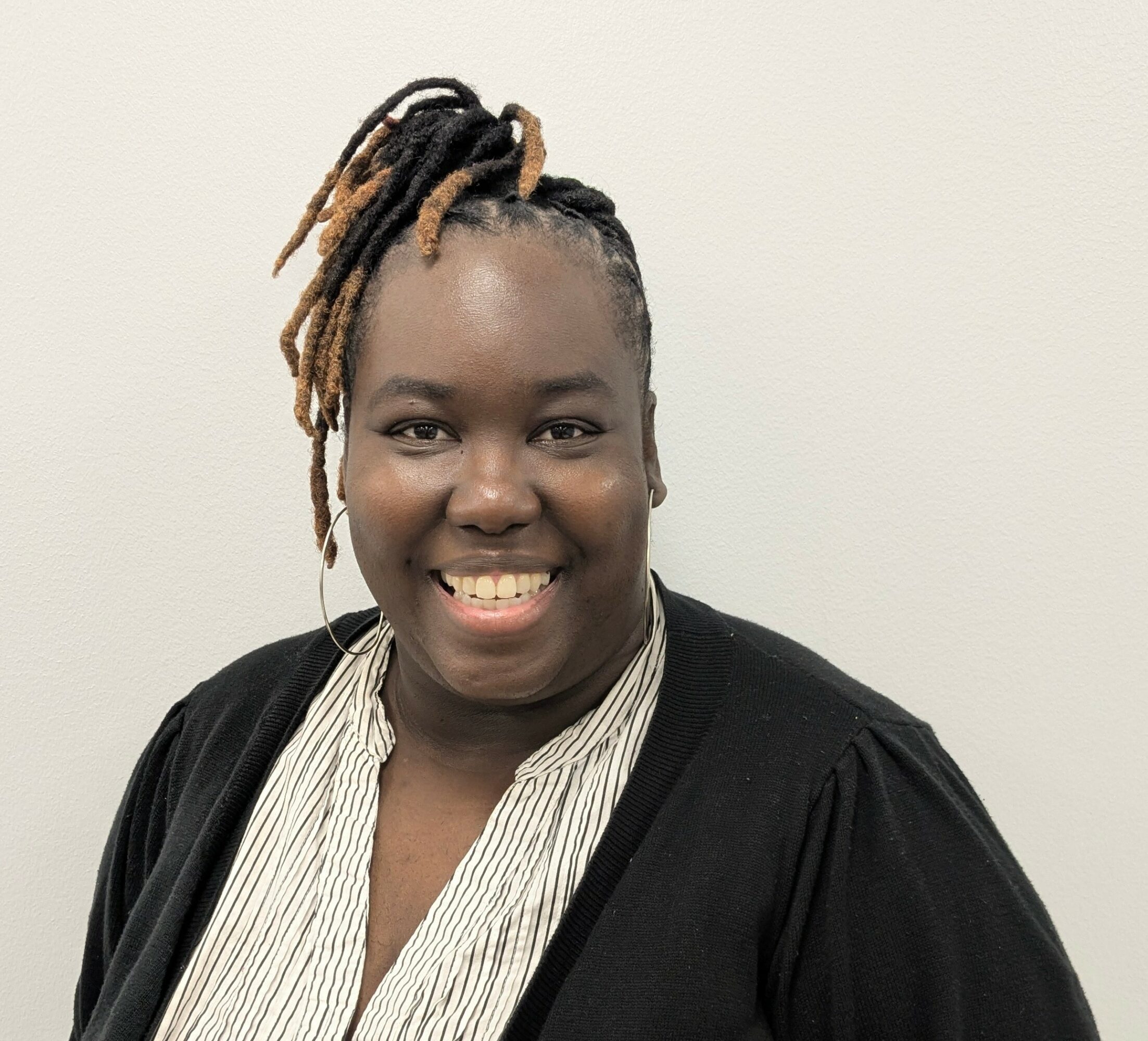 Smiling woman with dreadlocks styled in an updo, wearing a striped shirt and black cardigan, standing against a plain wall.